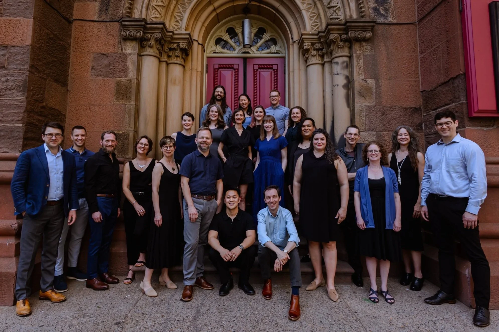 A group of singers in formal clothing in shades of black, blue, and gray, gathered outdoors on the steps to an ornate building. They look friendly and talented!