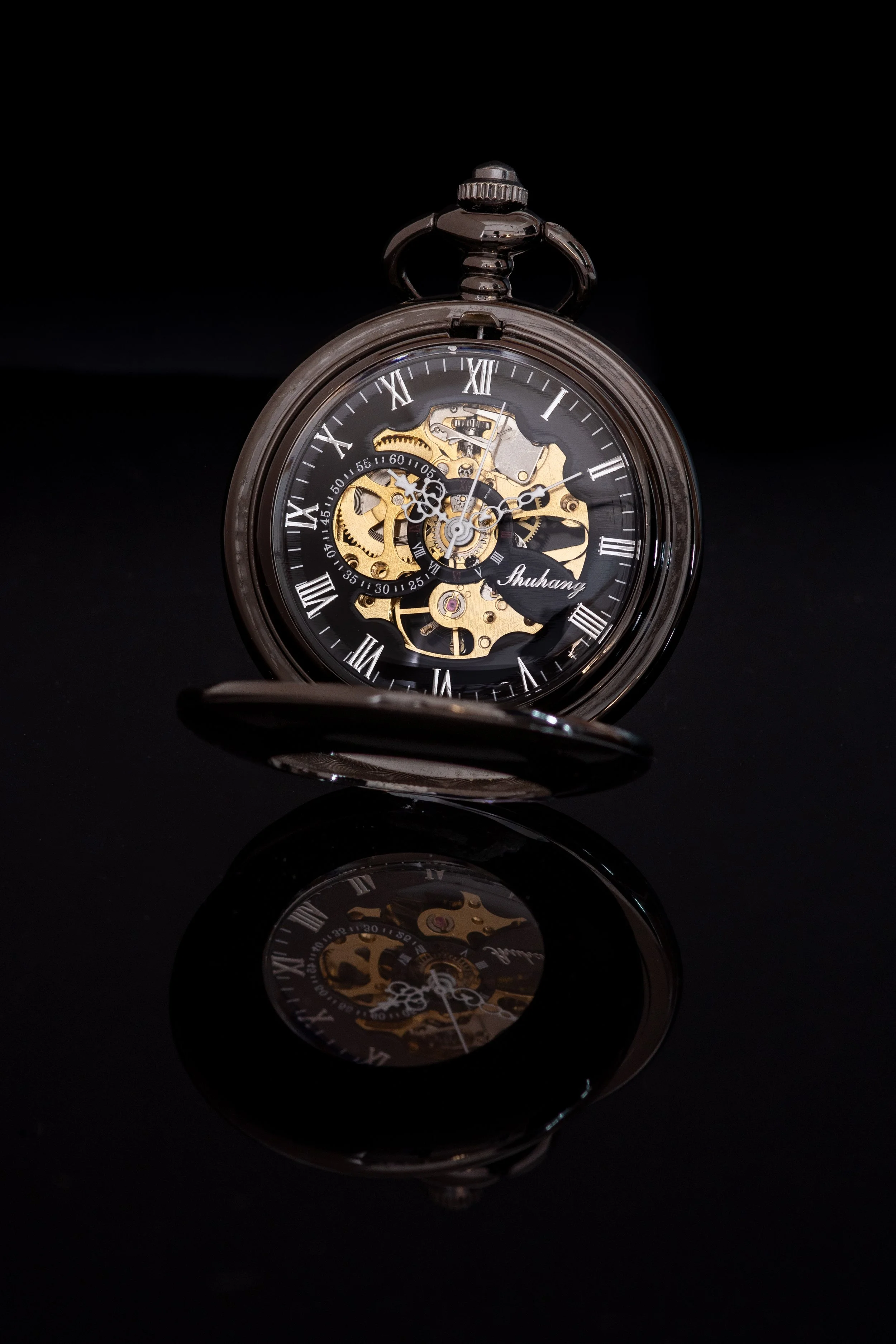Close-up of a vintage mechanical pocket watch with visible gears and Roman numerals on the black dial, set against a black background, with its reflection visible on a shiny surface.