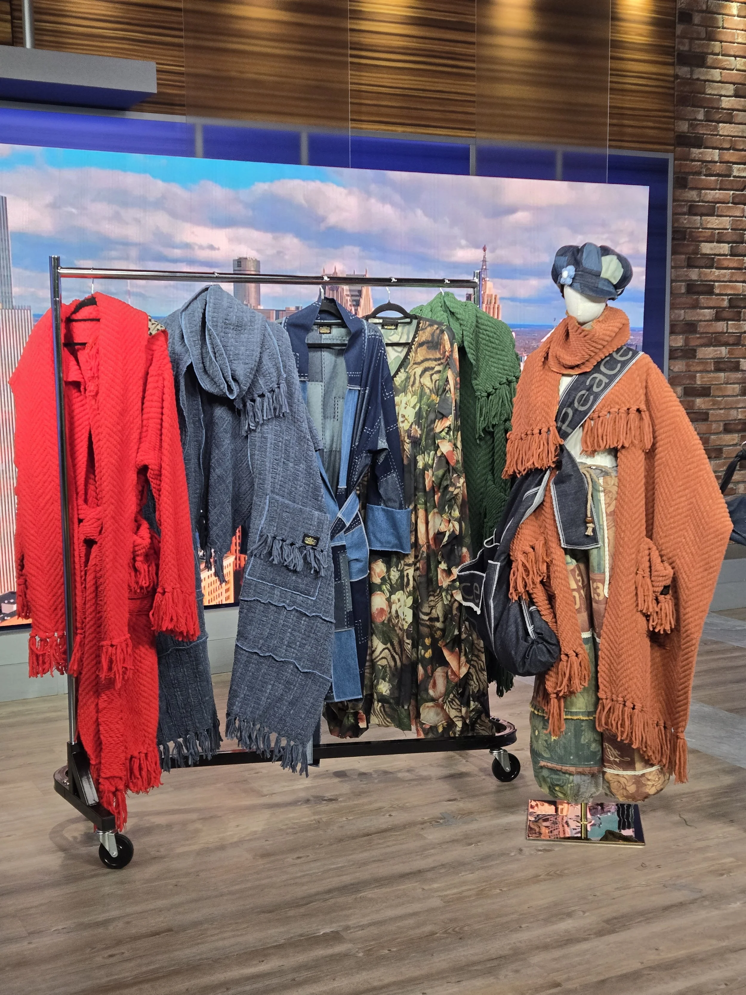Clothing display with various jackets and scarves, including red, gray, blue, green, and orange, on a rolling clothing rack and a mannequin with a denim patchwork hat, burnt orange scarf, and patchwork denim pants, against a cityscape backdrop.