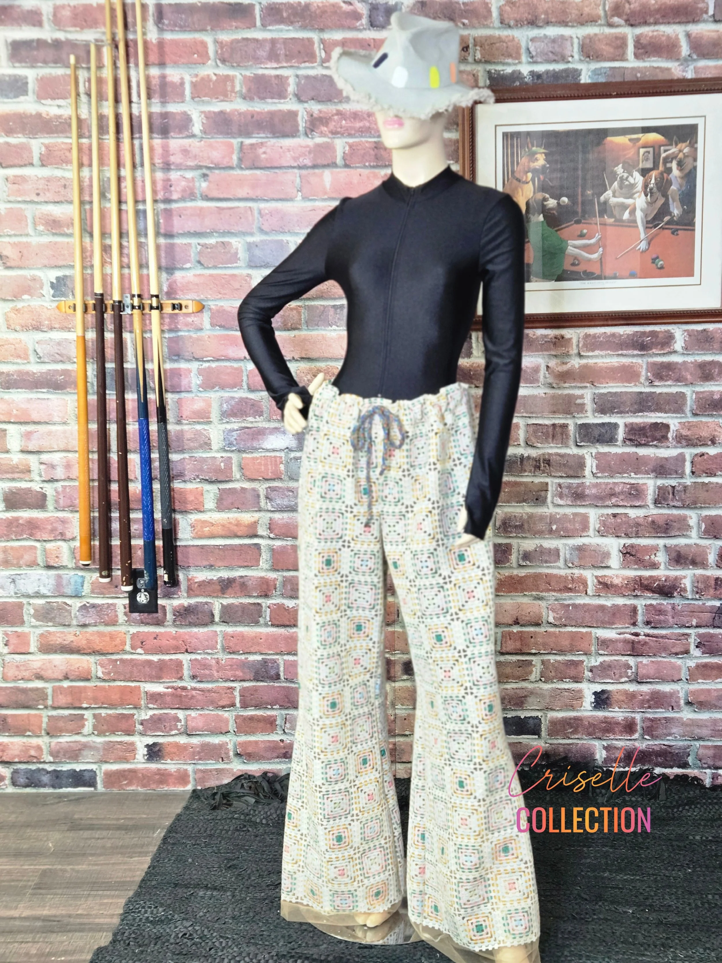 Mannequin dressed in a black long-sleeve top, patterned wide-leg pants, and a large, decorative hat, standing against a brick wall with hanging pool cues and a framed picture of dogs playing pool.