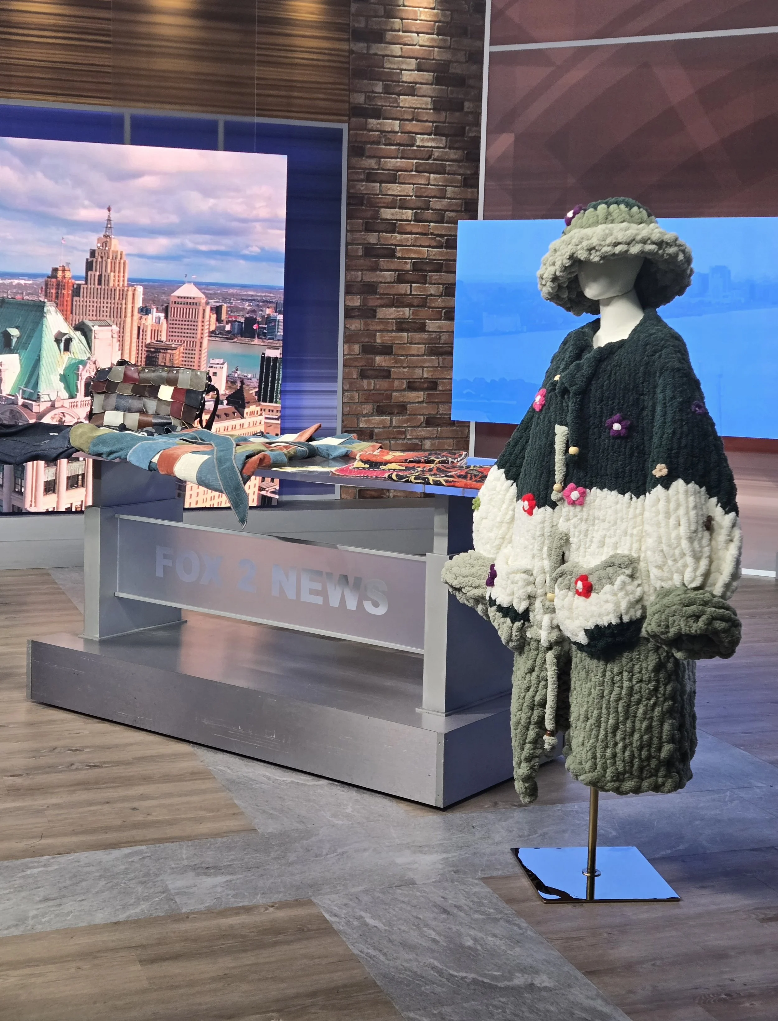 A display of handmade knitted clothing and accessories at a news station. Features a mannequin with a large, hat and matching coat, alongside a table with rolled-up fabric and a multicolored patchwork bag. There are cityscape photographs in the backg