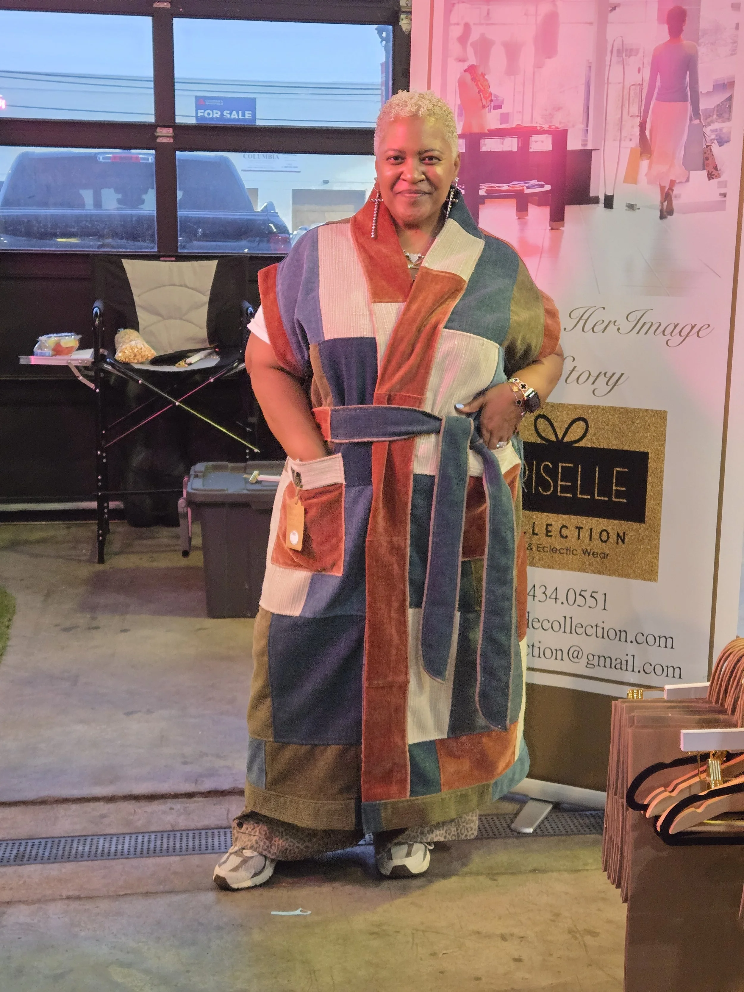 A woman standing indoors in front of a display banner and bay door, wearing a colorful custom made patchwork coat, with short blonde hair, smiling at the camera.