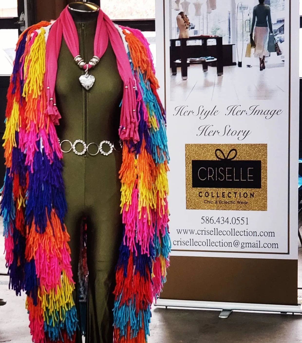 Colorfully decorated mannequin wearing a green outfit with a vibrant multicolored fringed shawl, pink scarf with a heart necklace, and a decorative belt, displayed next to a Criselle Collection sign with contact information.