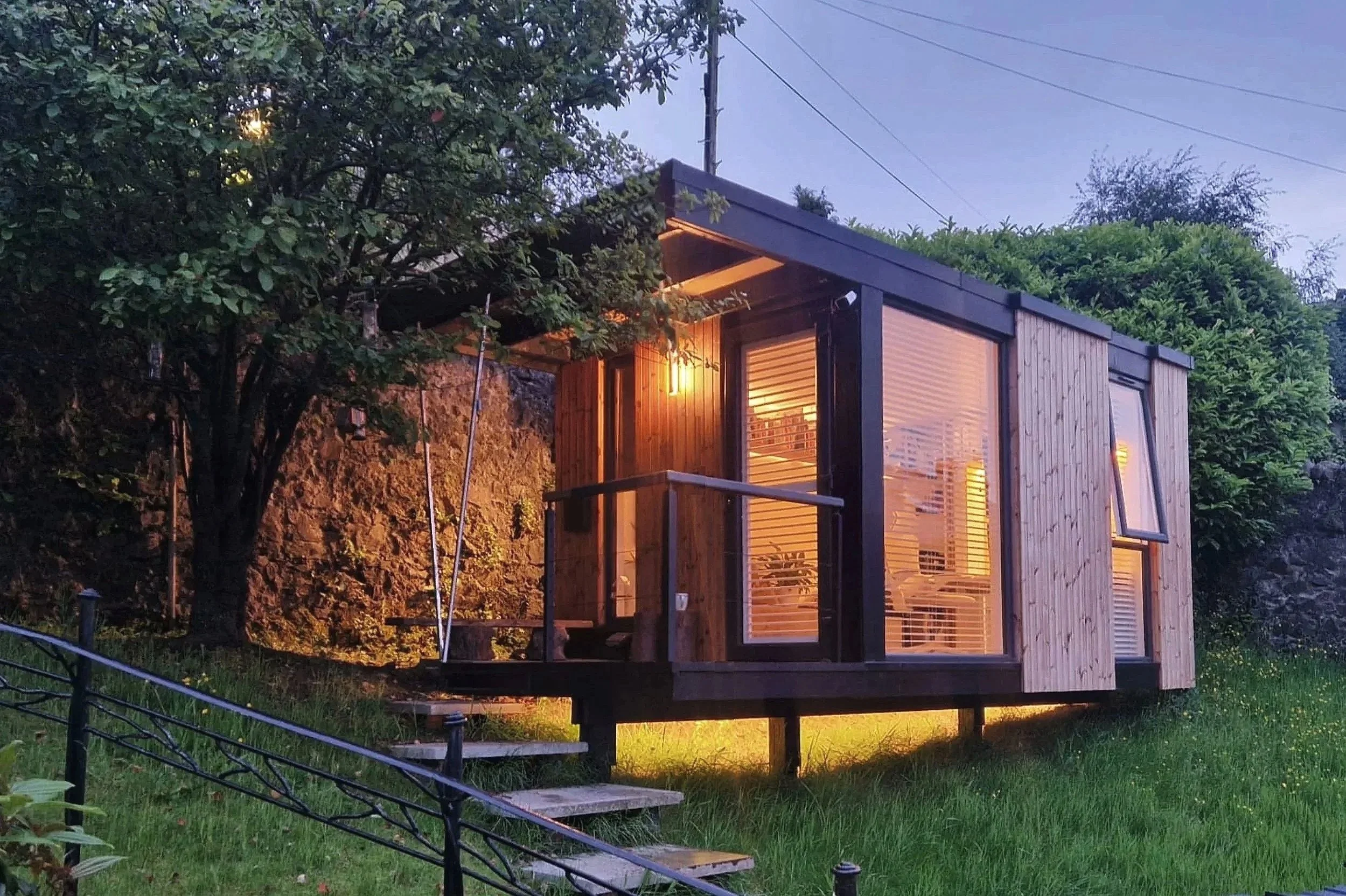 Modern tiny house with wood siding, large windows, and warm interior lighting, situated on a grassy hillside surrounded by trees, during dusk.
