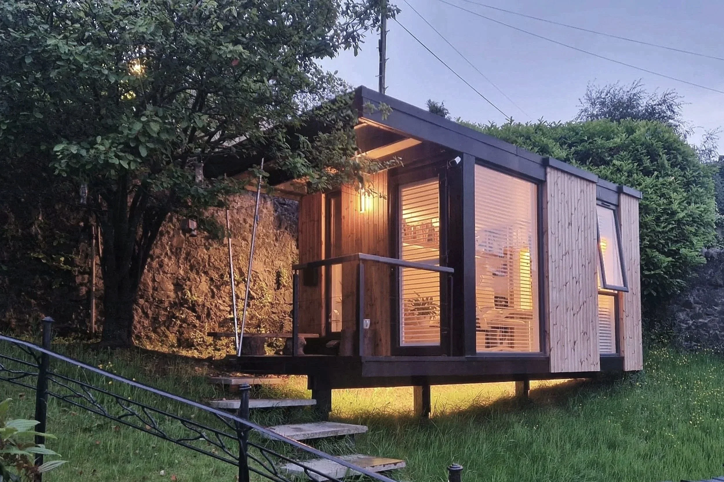 A modern Garden room  built with wood and glass, illuminated from inside, surrounded by trees and greenery, with a small staircase leading up to it at dusk. Langbank