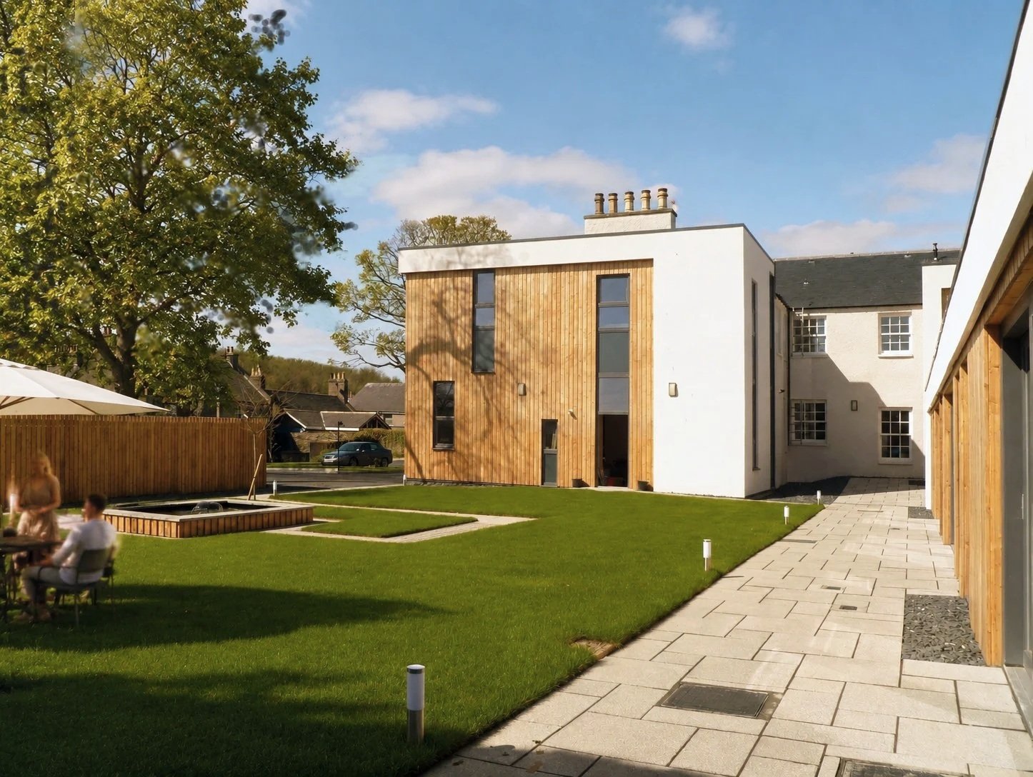 Keith lodge 12 unit flatted development .A modern residential building with a combination of white walls and wooden paneling, surrounded by a well-maintained lawn, paved walkways, and a large tree, with people sitting at a table outside.