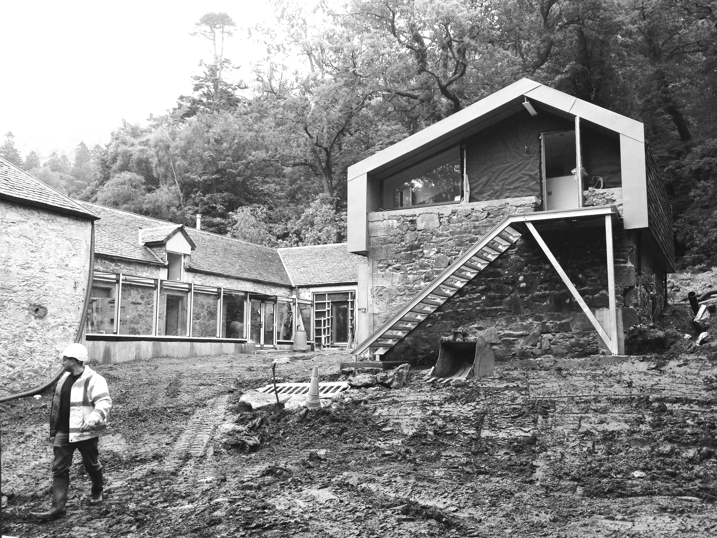 Construction workers on muddy ground outside a building under renovation, with a staircase and partially constructed walls, surrounded by trees.