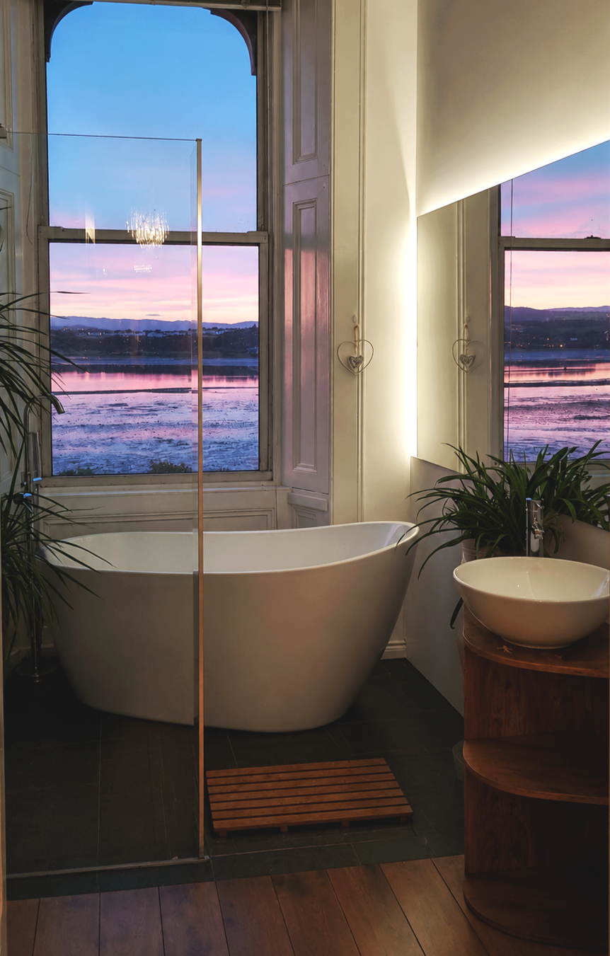 A modern bathroom with a white bathtub in front of large windows showing a colorful sunset over a lake or river. The bathroom has dark flooring, a wooden floor mat, a wooden stand with a ceramic basin sink, and potted plants.