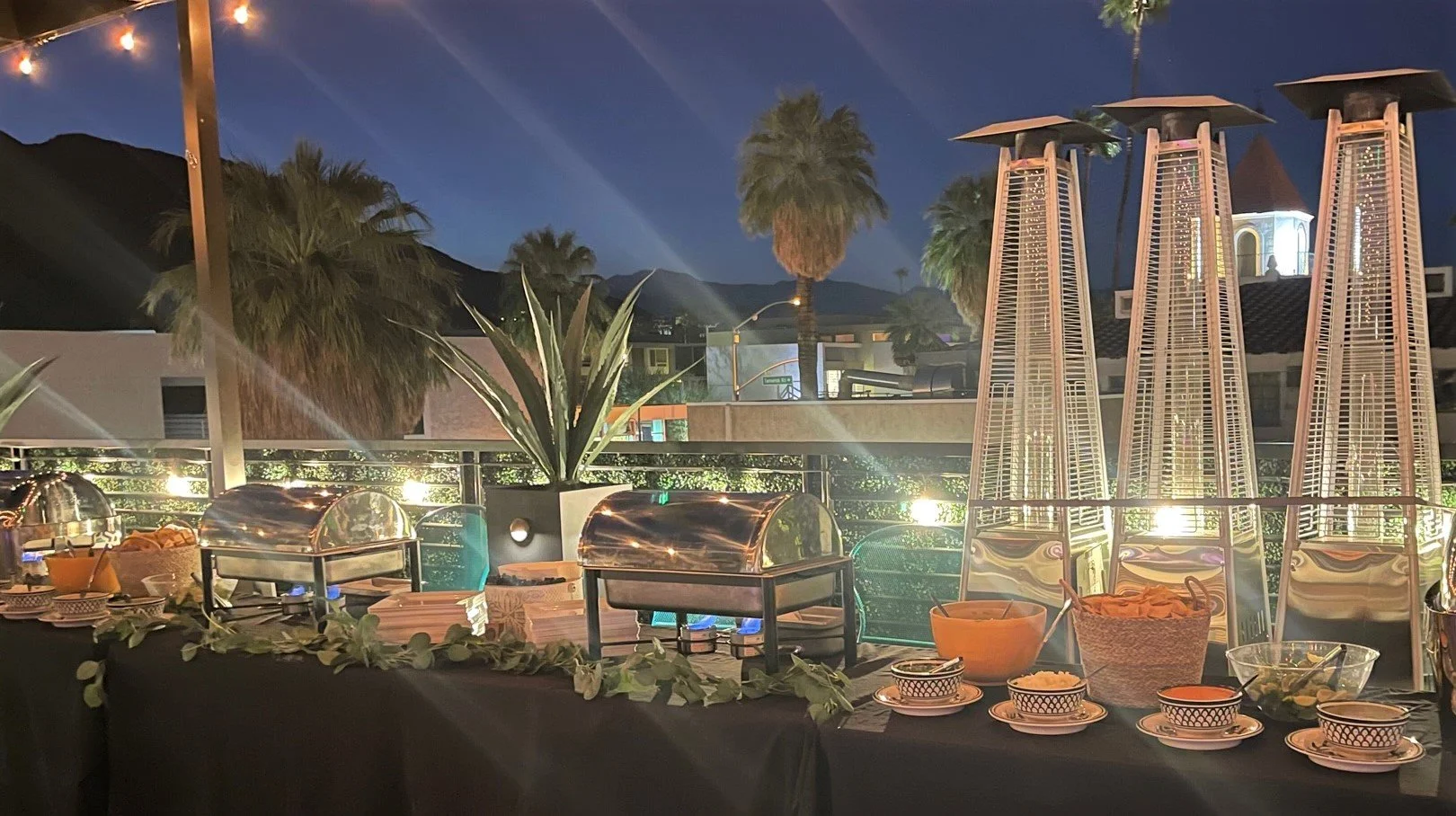 A full-service taco catering station on an outdoor terrace at night with stainless steel chafers, eucalyptus decor, and patio heaters in Orange County.
