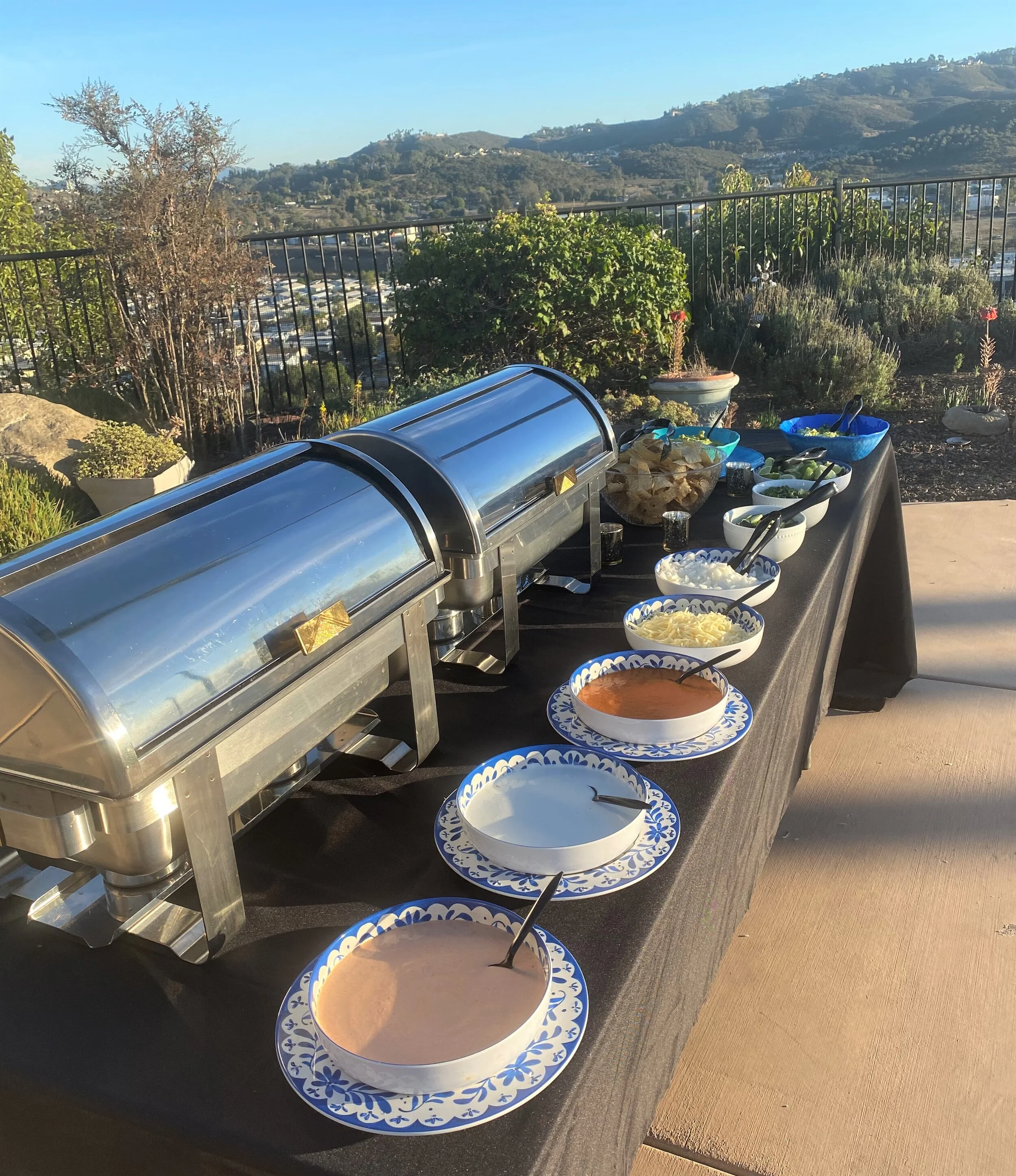 An elegant LaLa Tacos catering setup featuring polished stainless steel chafers and fresh artisan toppings in blue and white ceramic bowls, displayed on a black linen table overlooking a scenic Southern California valley.