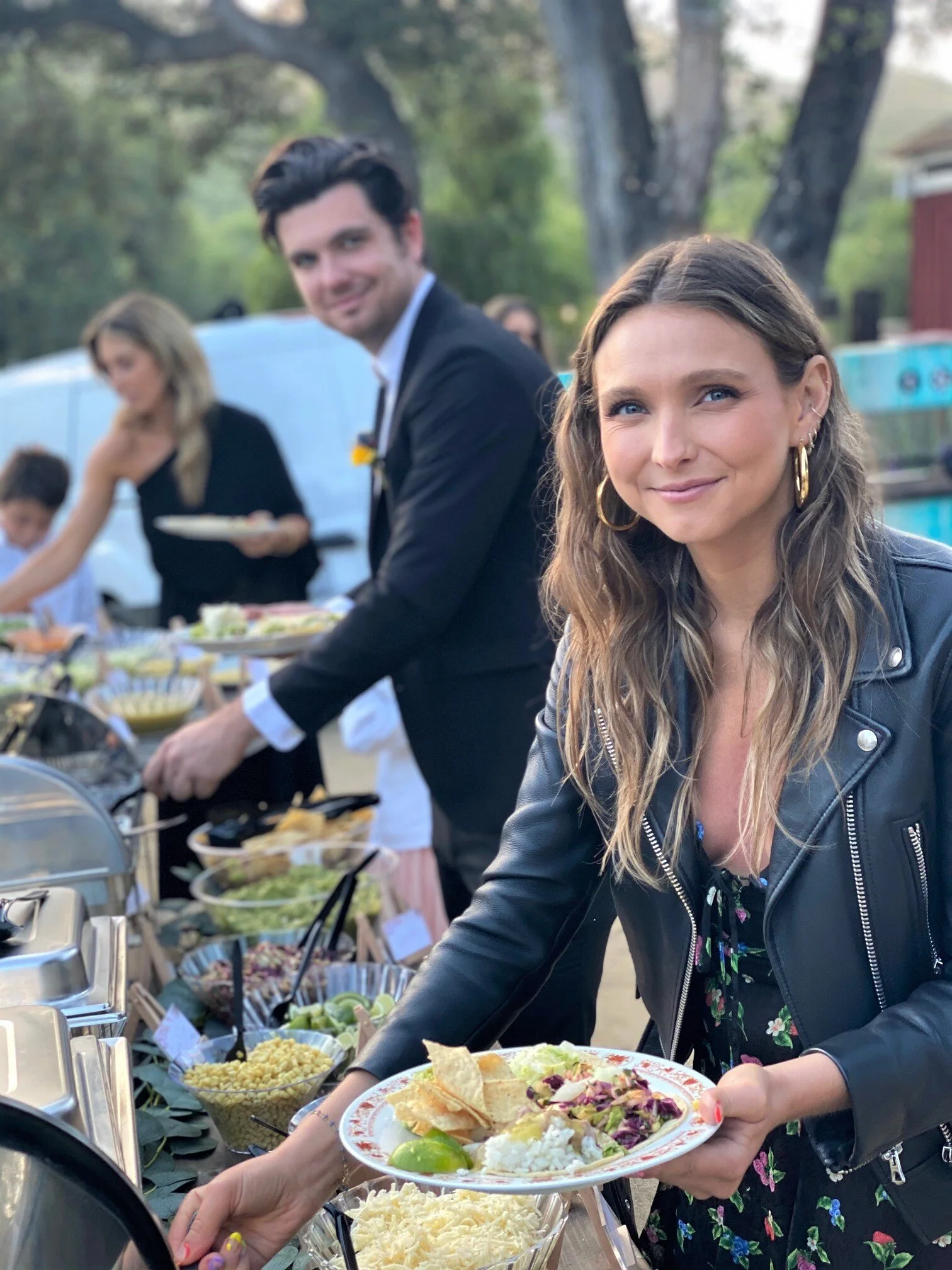 Smiling upscale wedding guests holding plates of food at an artisan outdoor taco bar buffet catering line