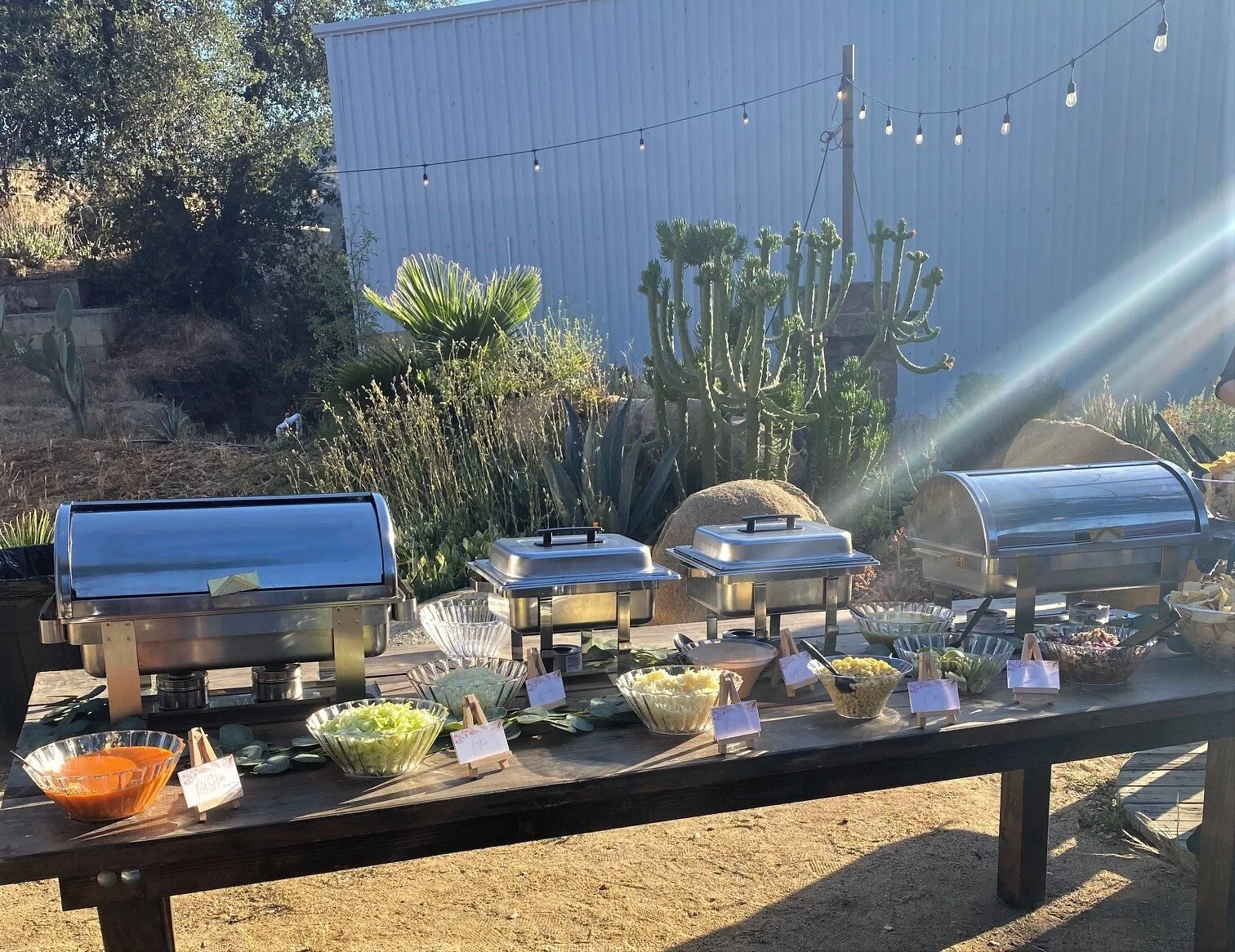 A premium LaLa Tacos catering setup on a rustic wooden table featuring professional stainless steel chafing dishes and fresh artisan toppings in glass bowls for a San Diego outdoor wedding.