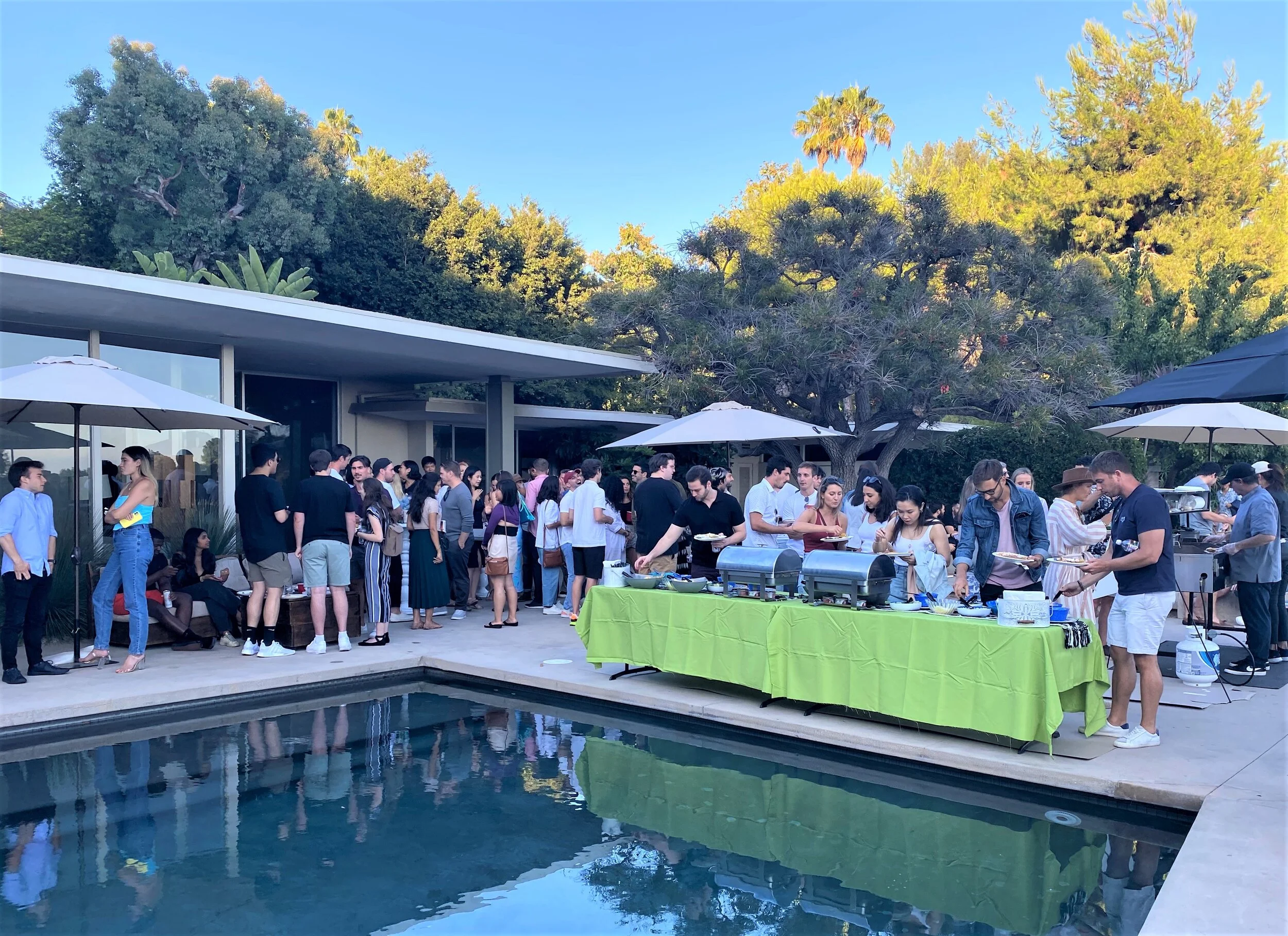 A large crowd of guests enjoying a premium poolside taco bar featuring professional stainless steel chafers and a live-action cooking station at a luxury mid-century modern estate in Beverly Hills.