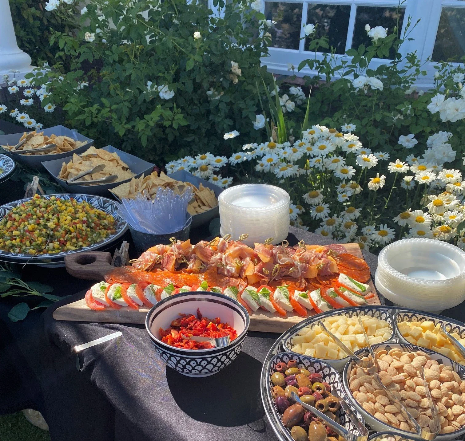 An elegant outdoor wedding appetizer grazing table by LaLa Tacos featuring a wooden charcuterie board with caprese salad and artisan skewers surrounded by fresh salsas and gourmet cheeses next to blooming white flowers.