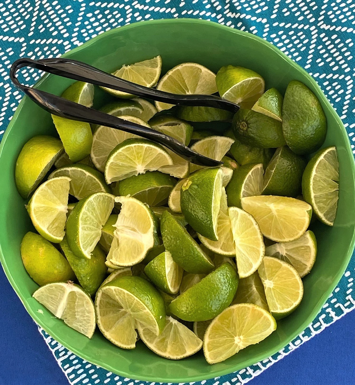 A bright green bowl filled with freshly sliced crisp lime wedges and black serving tongs on a vibrant turquoise tablecloth