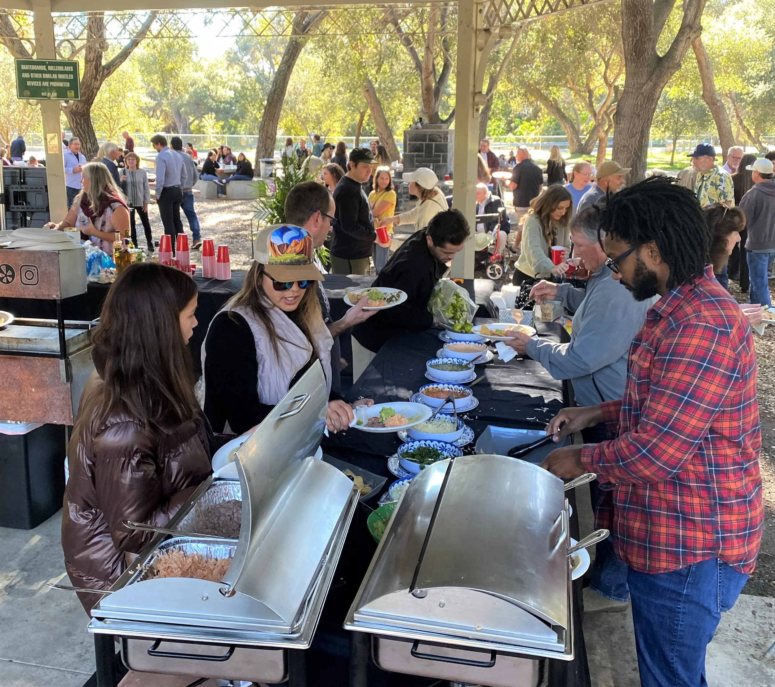 A professional boutique Mexican catering setup at a park pavilion for a celebration of life. Guests are being served from sleek stainless steel chafing dishes on a black-linened buffet line, featuring authentic tacos and scratch-made salsas.
