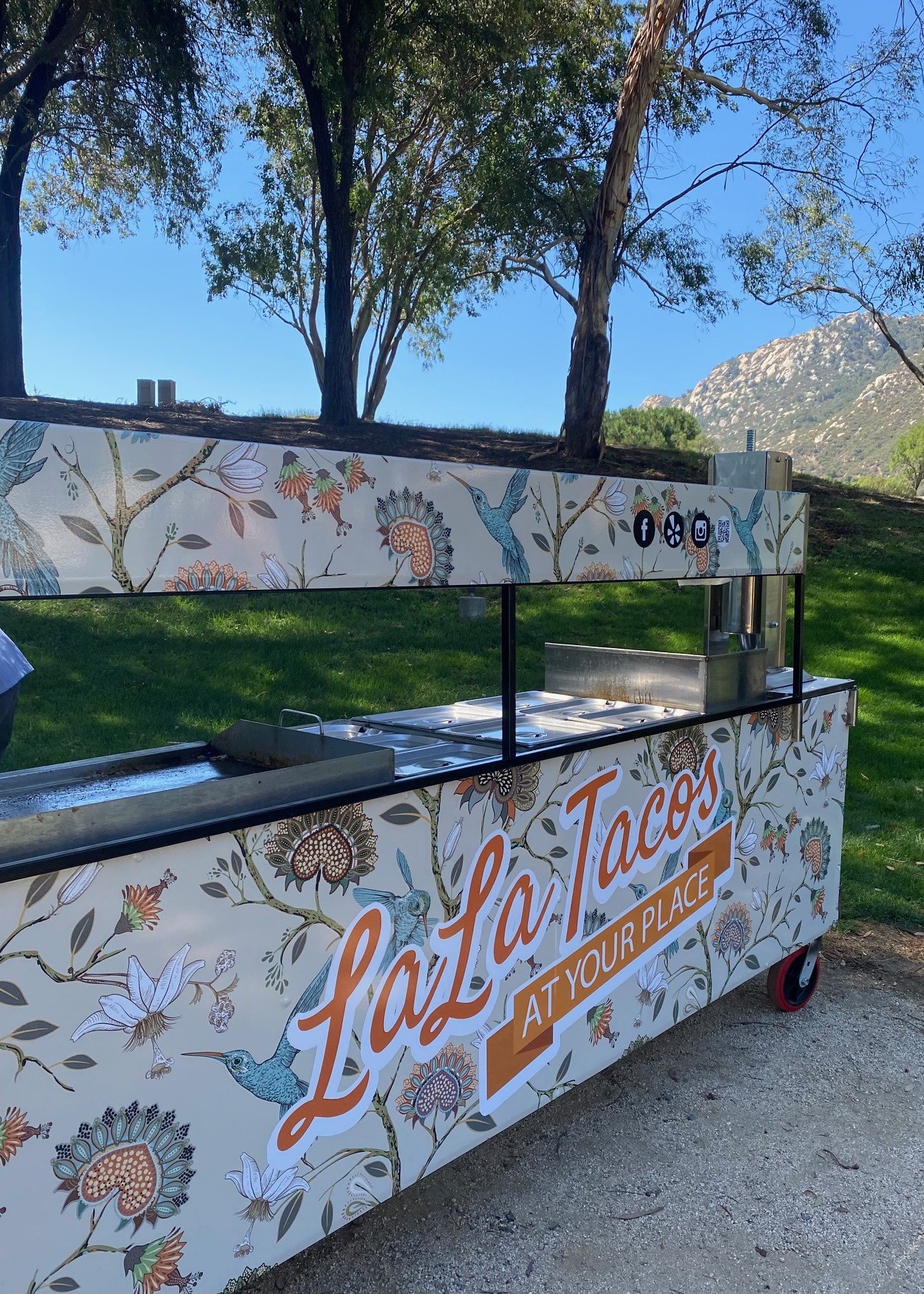A boutique, custom-wrapped LaLa Tacos mobile catering cart featuring a high-heat plancha and integrated stainless steel warming pans, showcasing a clean, self-contained artisan setup at a scenic outdoor venue.