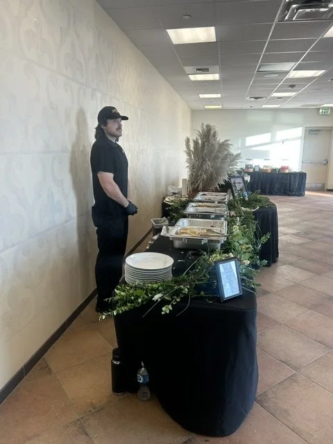 Uniformed LaLa Tacos professional ready to serve guests at an elegant indoor taco buffet station featuring black linens, fresh greenery, and white dinner plates.
