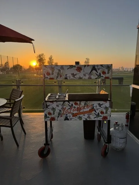 Custom hummingbird-wrapped LaLa Tacos catering cart set up on a glass balcony overlooking a golf course at sunset in Orange County.