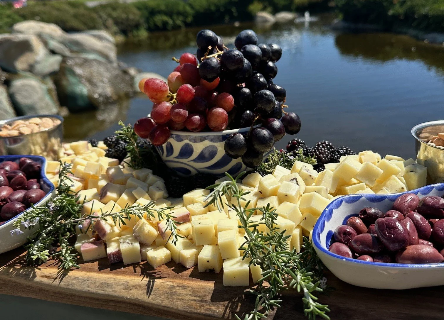 Artisan charcuterie grazing table with cubed gourmet cheeses fresh grapes and kalamata olives on a wooden board overlooking a scenic lake