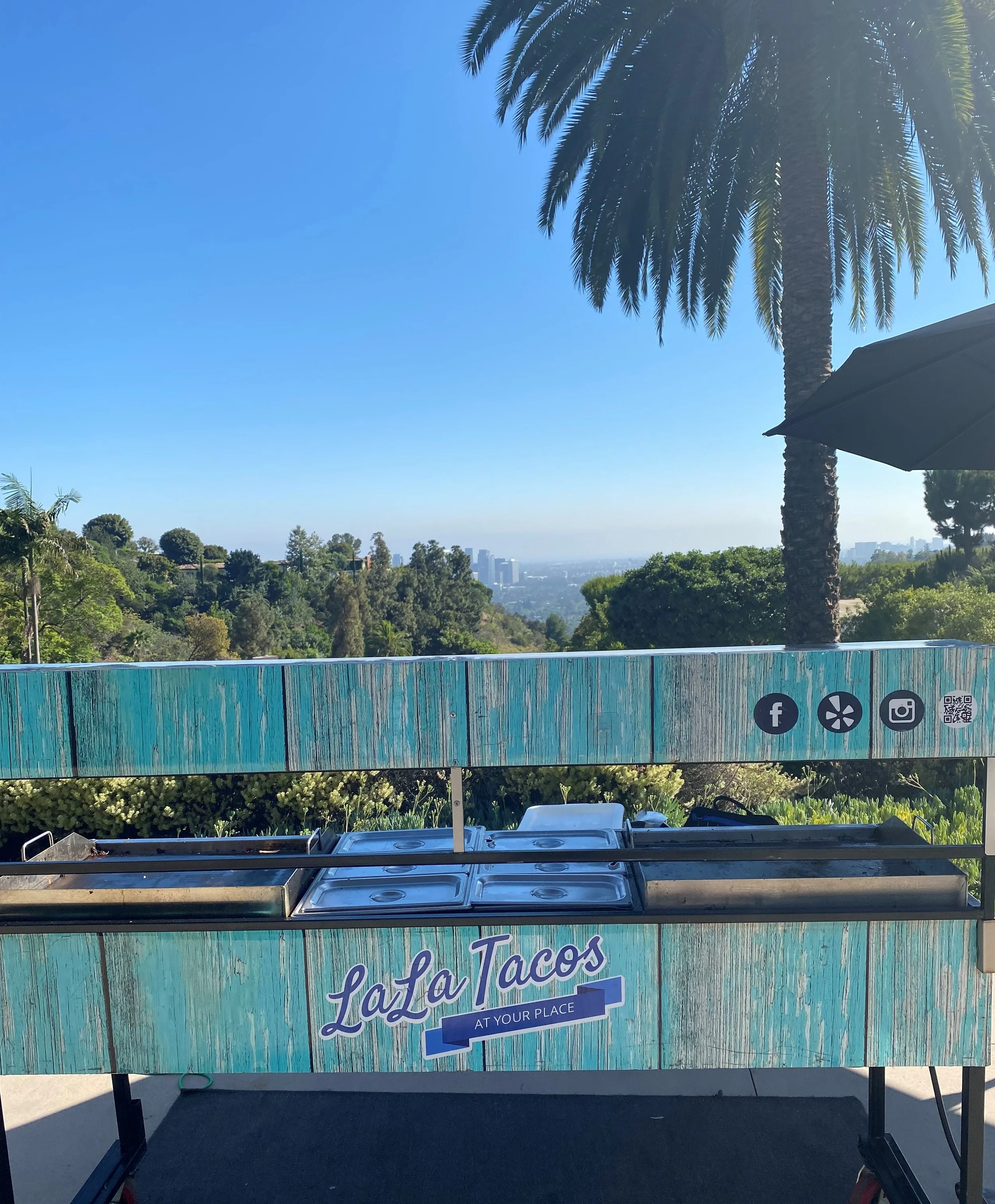 A custom-wrapped LaLa Tacos live-action catering cart featuring high-heat planchas, set up at a luxury private residence in Beverly Hills with the downtown Los Angeles skyline in the background.