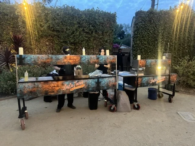 Custom patina-wrapped LaLa Tacos catering cart and matching churro station deployed for a large-scale 575-guest event against a lush greenery wall in San Diego, California.
