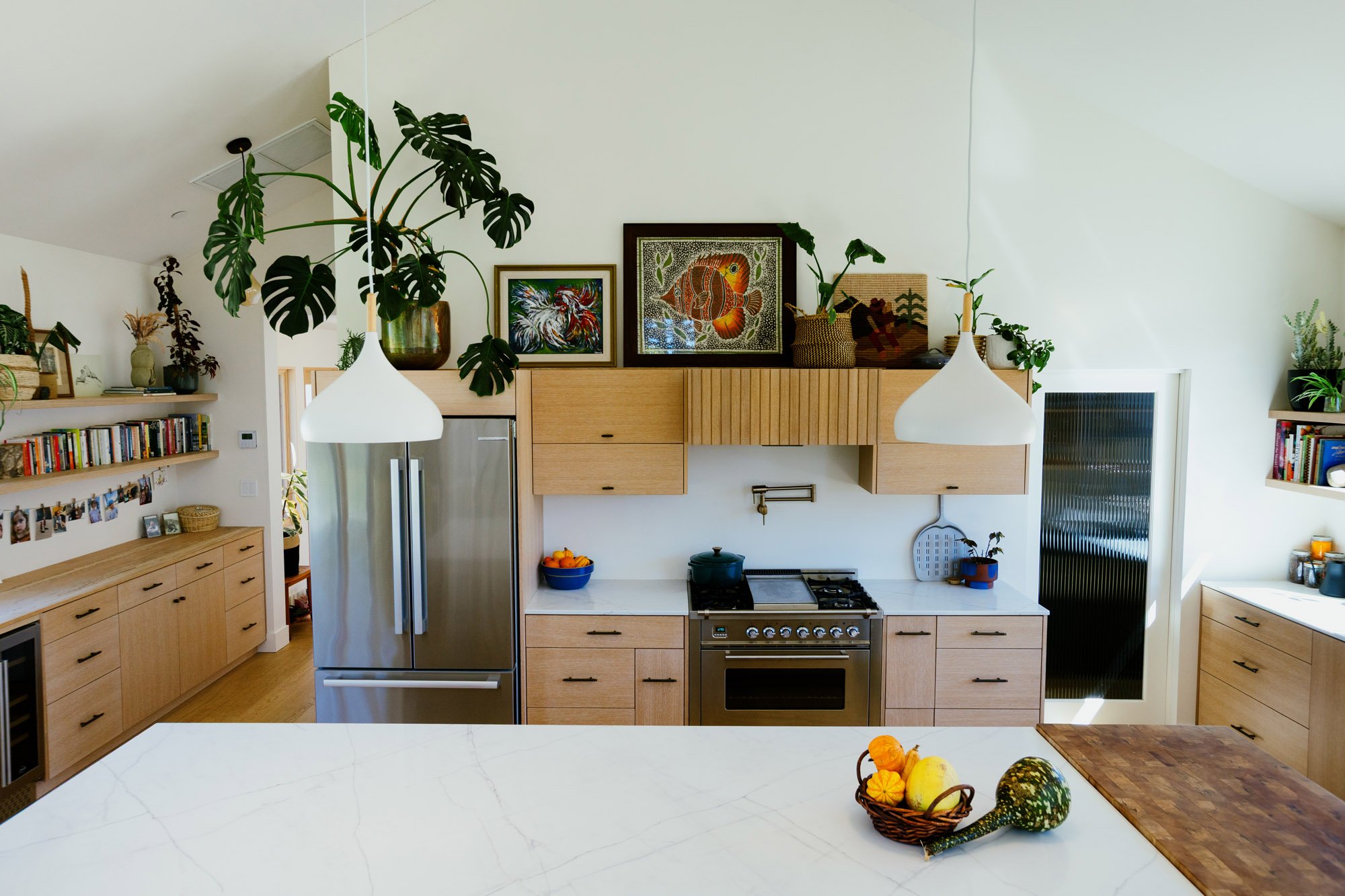 A modern kitchen with light wood cabinets, a stainless steel refrigerator, a gas stove, and a white marble kitchen island with a basket of decorative gourds and pumpkins.
