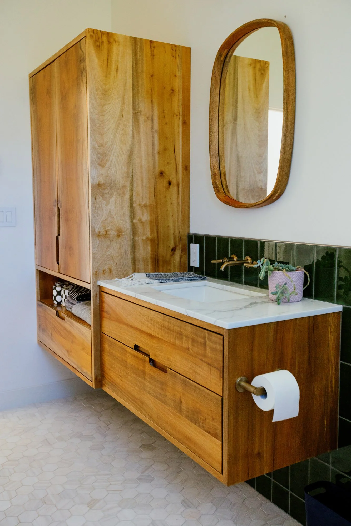 A modern bathroom vanity made of wood with a white marble countertop, a built-in sink, and a brass faucet. There is a small potted plant and a folded towel on the countertop. A frameless oval mirror is mounted above the vanity, and a green tiled back