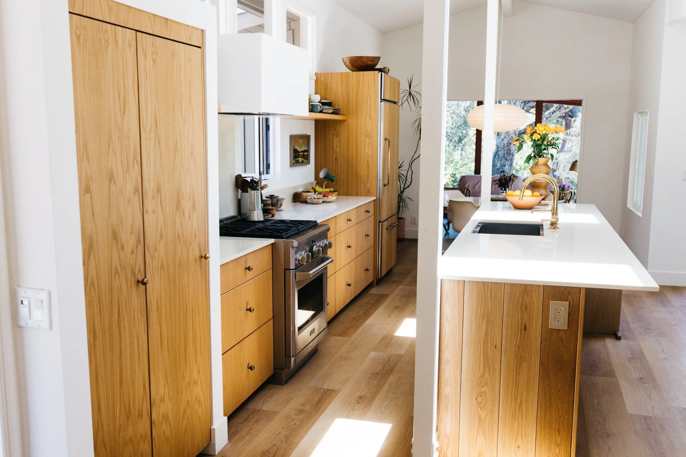 A bright, modern kitchen with wooden cabinets, a white countertop, and a stainless steel stove. There are large windows and a view of trees outside, with decorative plants and bowls on the counter.