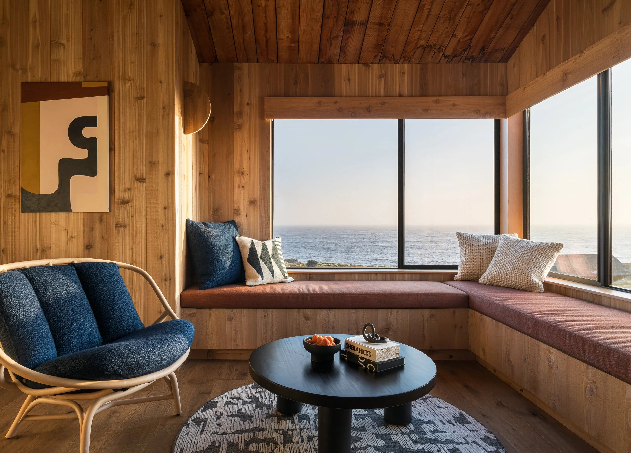 A cozy living room with wooden interior walls and ceiling, a large window showing an ocean view, a blue armchair, a built-in window seat with cushions, a round black coffee table with a bowl of snacks and books, and an abstract painting on the wall.