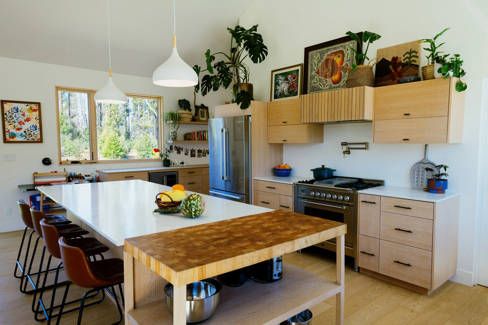 A modern kitchen with wooden cabinets, white countertops, and stainless steel appliances. A large white kitchen island with chairs around it, topped with decorative gourds. There are large windows overlooking a wooded outdoor area, and the walls are 