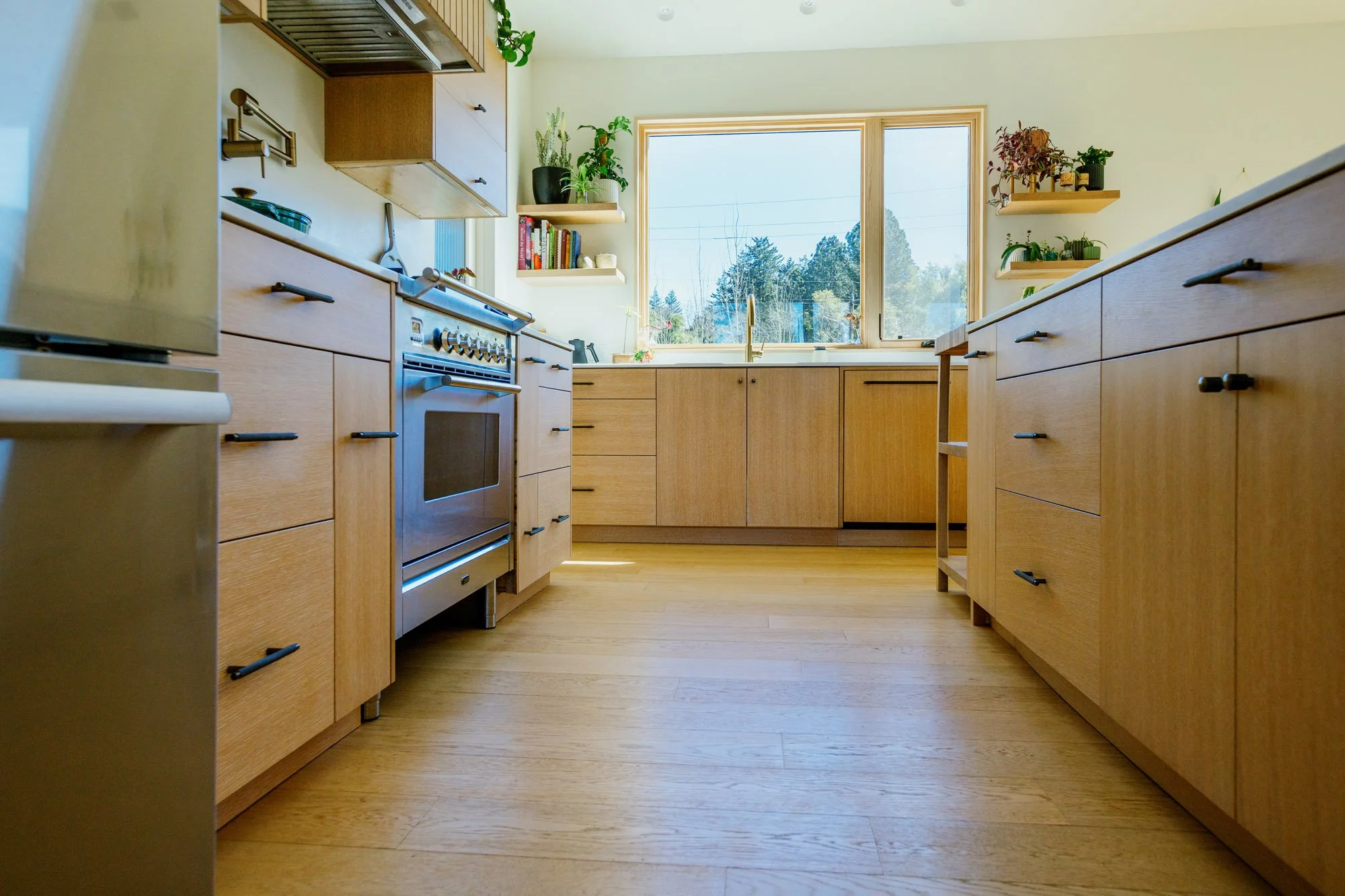 A bright kitchen with light wooden cabinets and flooring, a large window showing trees outside, and plants on shelves.