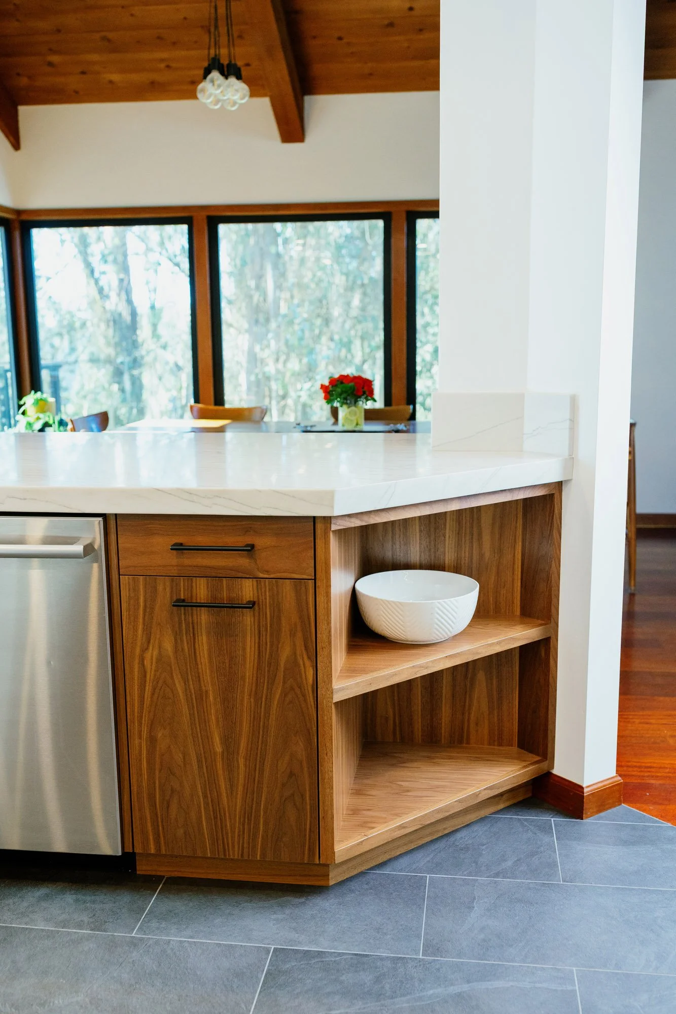 Close-up of a kitchen island with a white marble countertop and wooden cabinetry, with a view of a bright room with large windows and a red flower arrangement in the background.