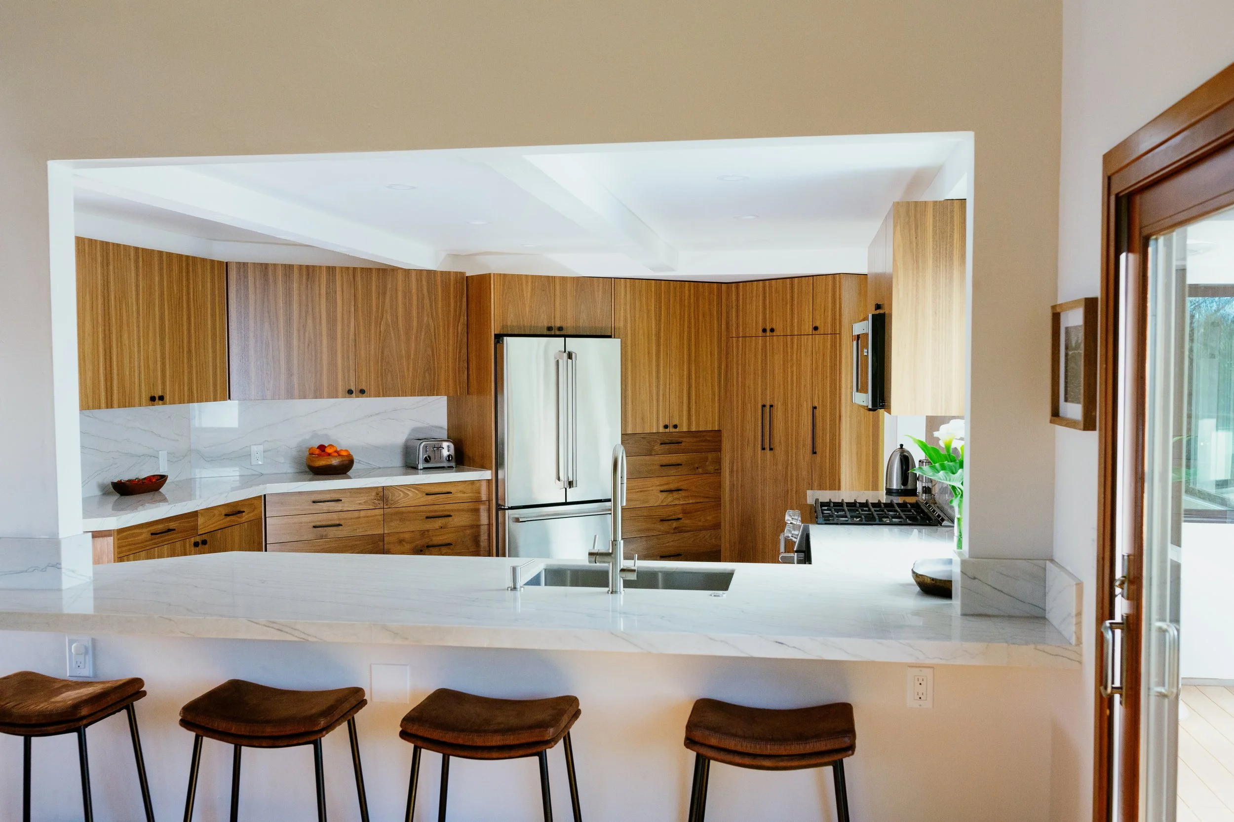Modern kitchen with wooden cabinets, marble countertops, and white walls, featuring a large window on the right and a breakfast bar with four brown stools in the foreground.