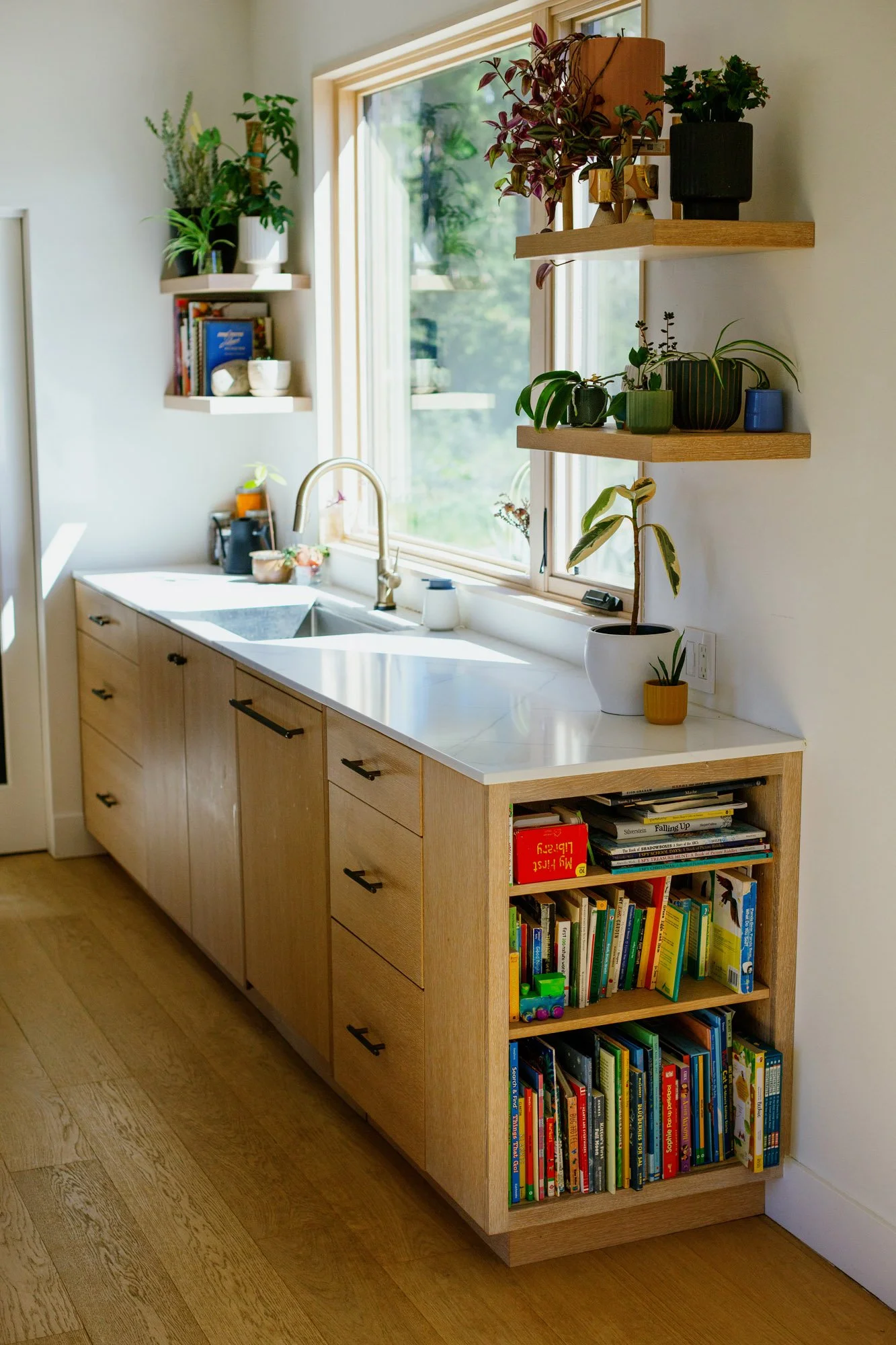 A kitchen with a wooden cabinet that has books and a kitchen sink with a window above it. Two floating shelves hold potted plants and books. The room is bright with sunlight.