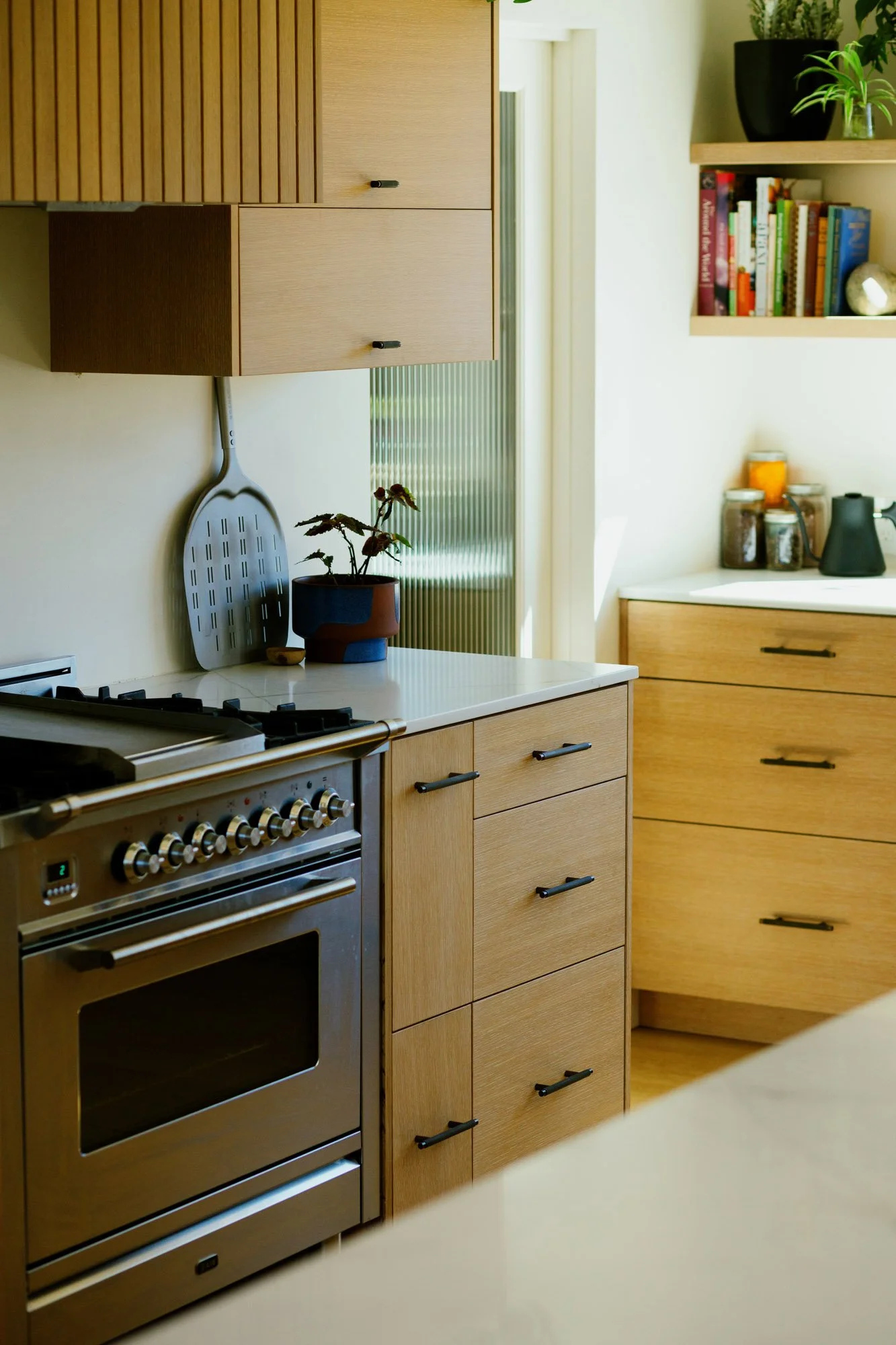 A modern kitchen with wooden cabinets, a stainless steel oven, a pot with a plant, and a shelf with books and jars.