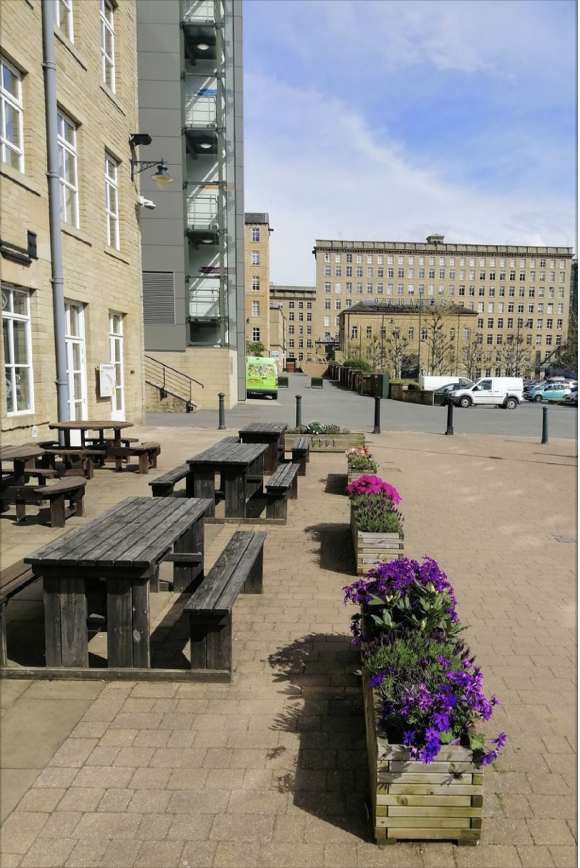 Dean Clough Mills in Halifax , looking down on the buildings from the car park outside. Wooden tables and benches and planters with purple flowers in the bottom half of. the photo.