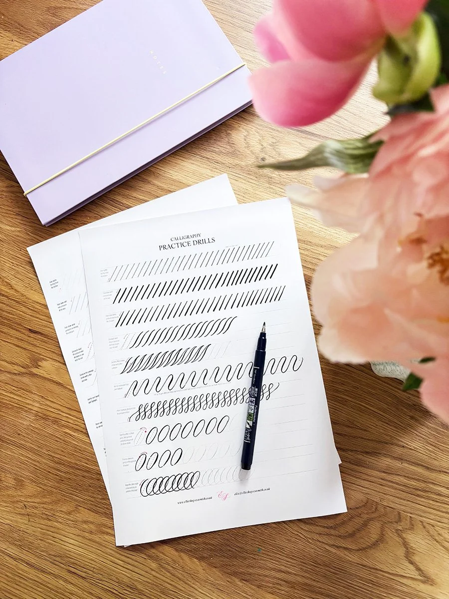a table with calligraphy drills worksheet and peony flowers in a vase and notebook and brush pen