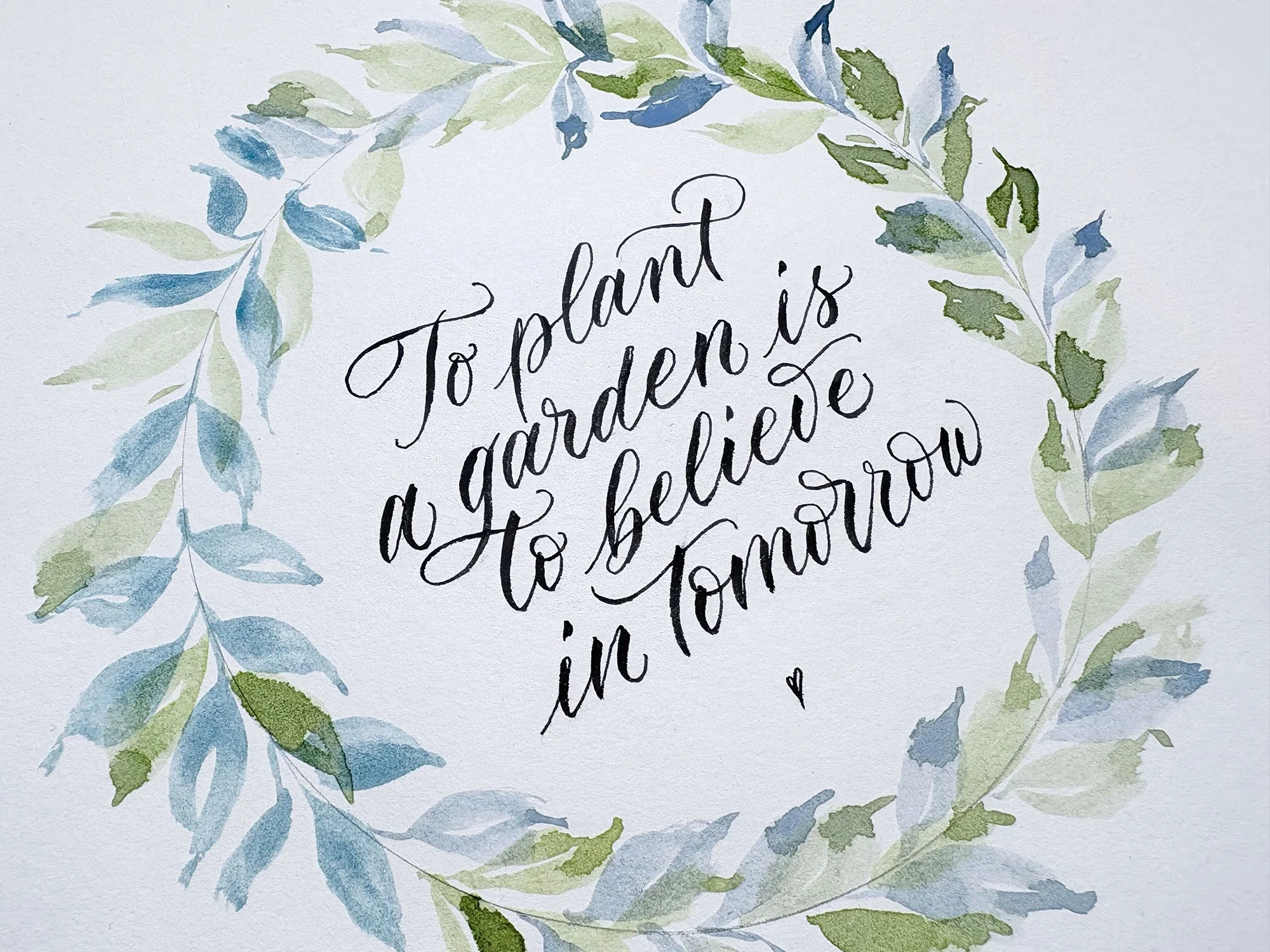 A quote written with a brush pen surrounded by a wreath of blue and green watercolour leaves.