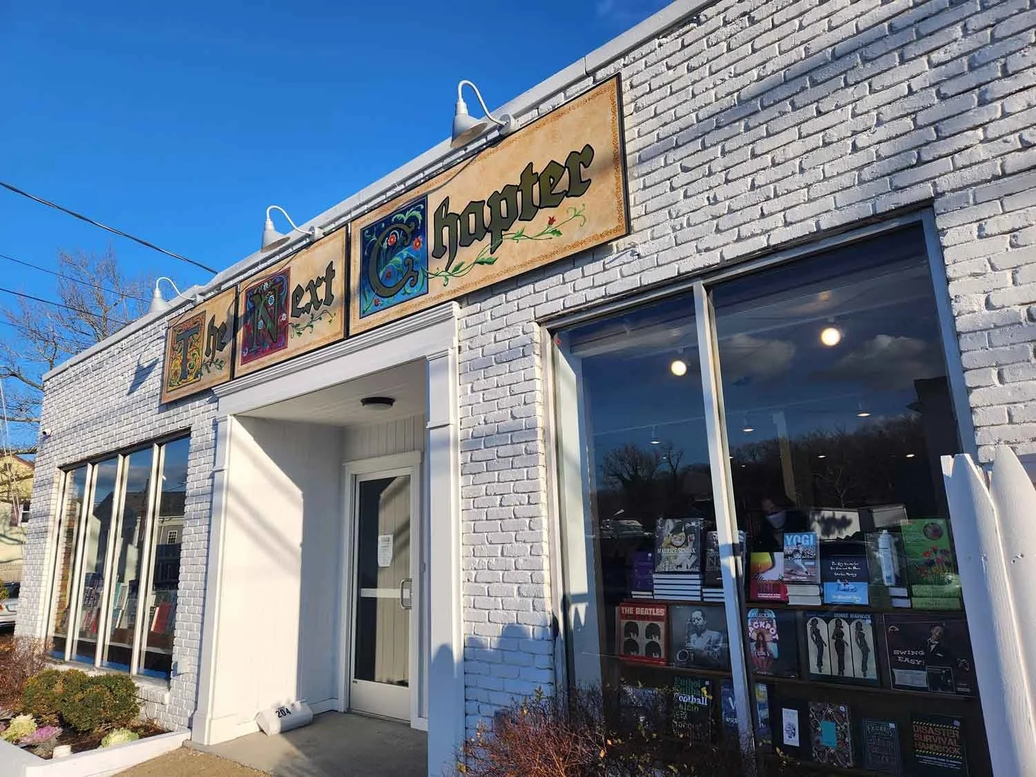A photo of a bookstore called The Next Chapter Bookstore with a white brick exterior and large windows.