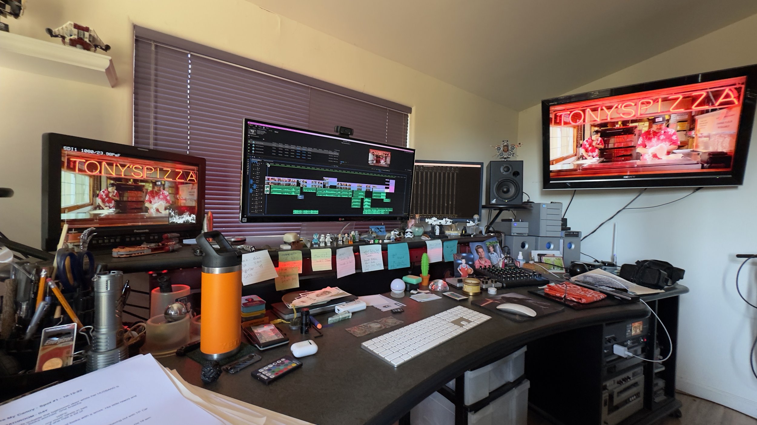 A cluttered desk at Studio 8 with three monitors, one displaying a video editing software, another showing a video of a pizzeria called Tony's Pizza, and the third with a recording or editing program.