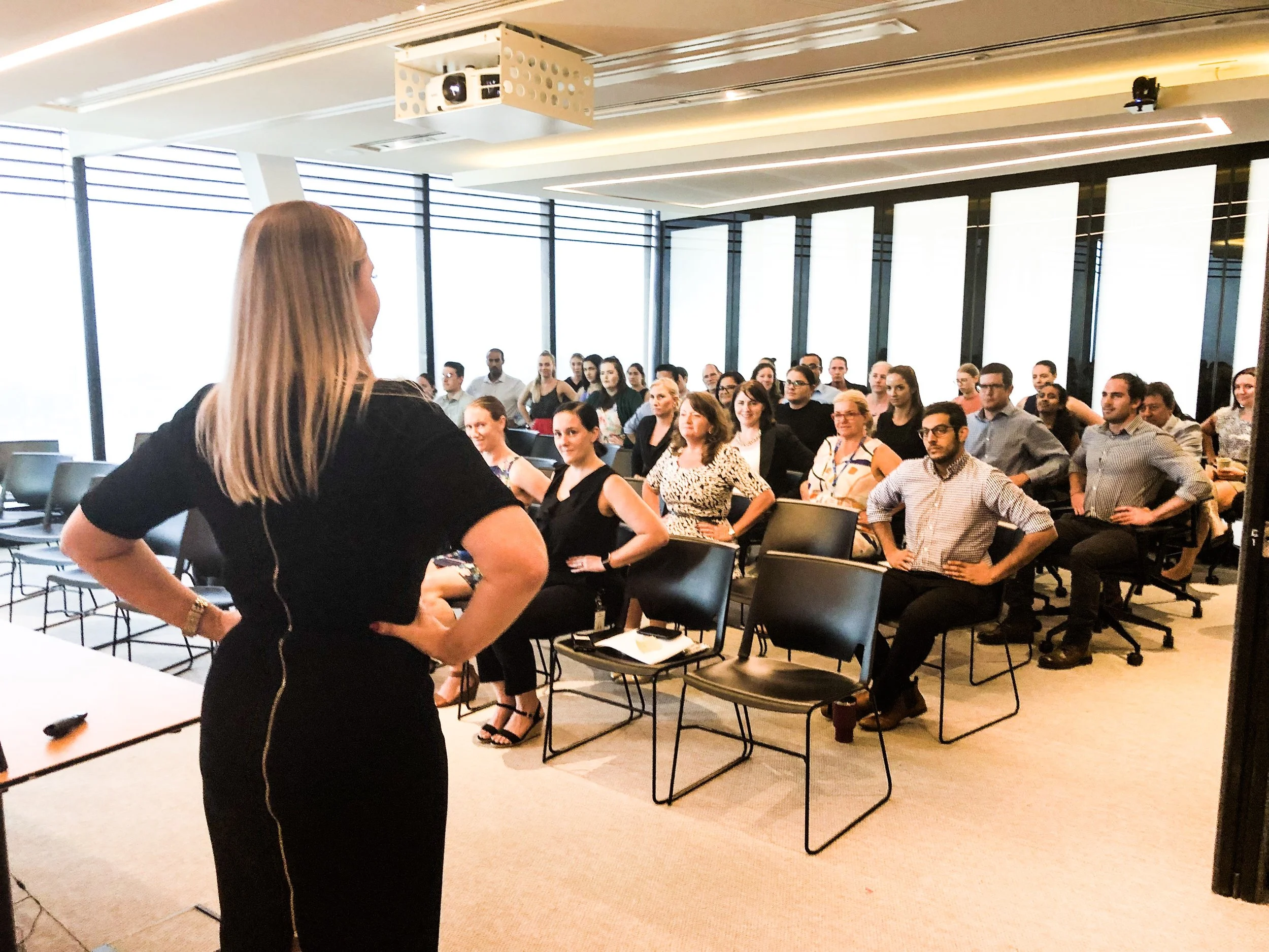 Sally Prosser team workshop, standing in front of a room full of seated people, giving a presentation or speech in a modern office conference room with large windows.