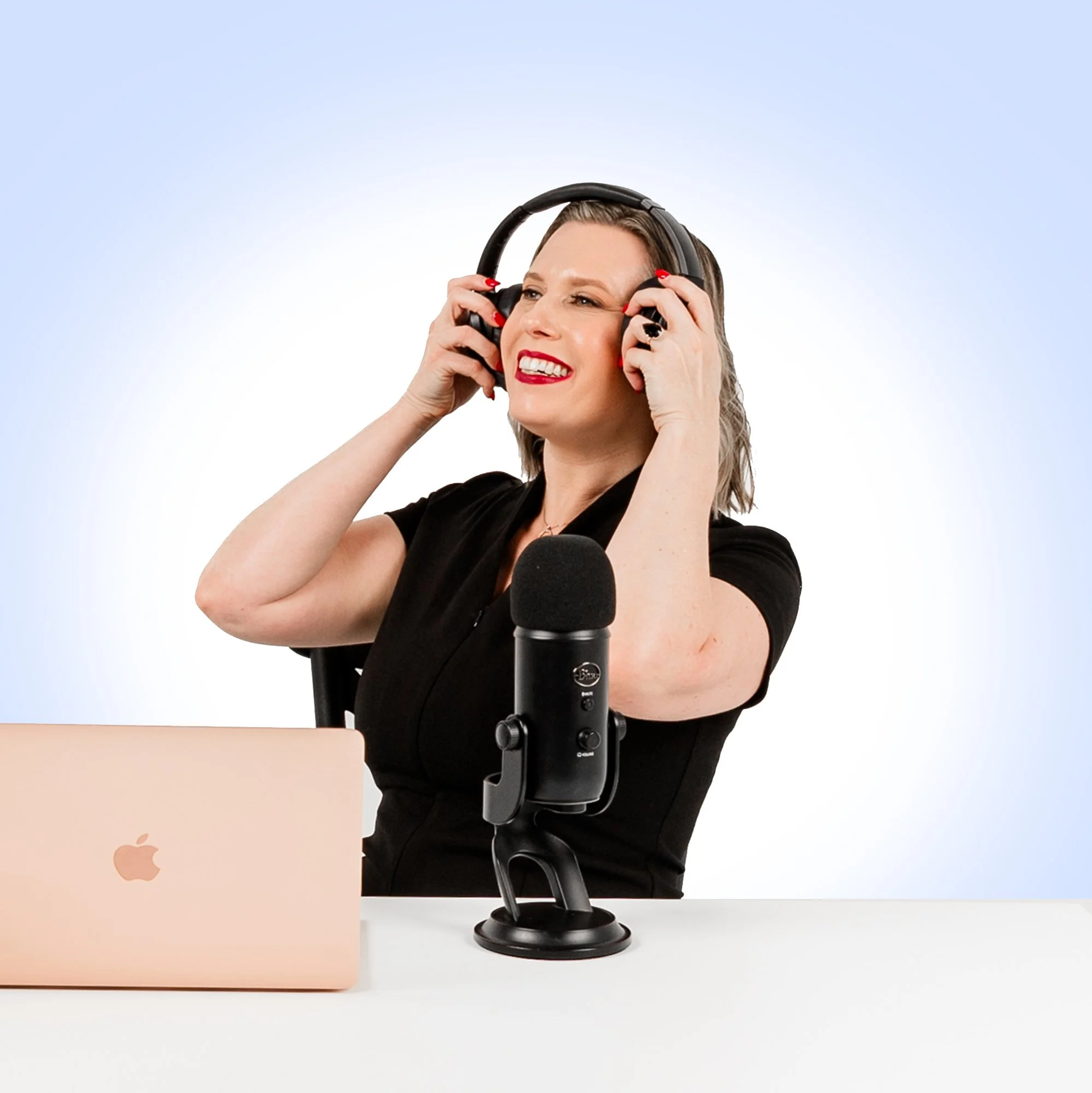 Sally Prosser, wearing black shirt with headphones, sitting at desk with a pink Apple MacBook and a microphone in front, grimacing or singing.