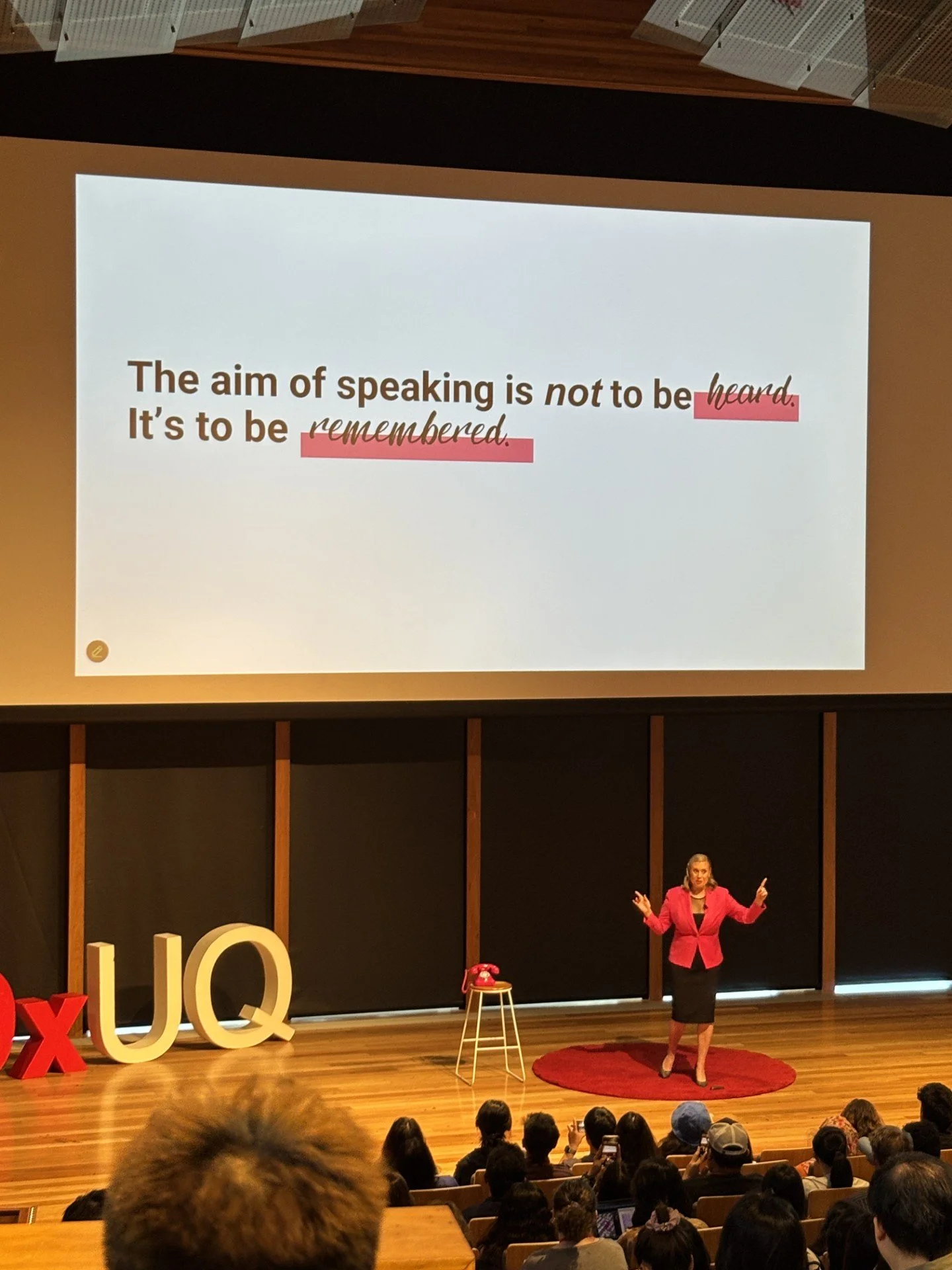 a pink blazer and black skirt giving a presentation on stage in front of an audience, with a large screen behind her displaying a slide with the text: "The aim of speaking is not to be heard. It's to be remembered." Large decorative letters "UQ" are on the stage. The audience is seated and watching attentively.