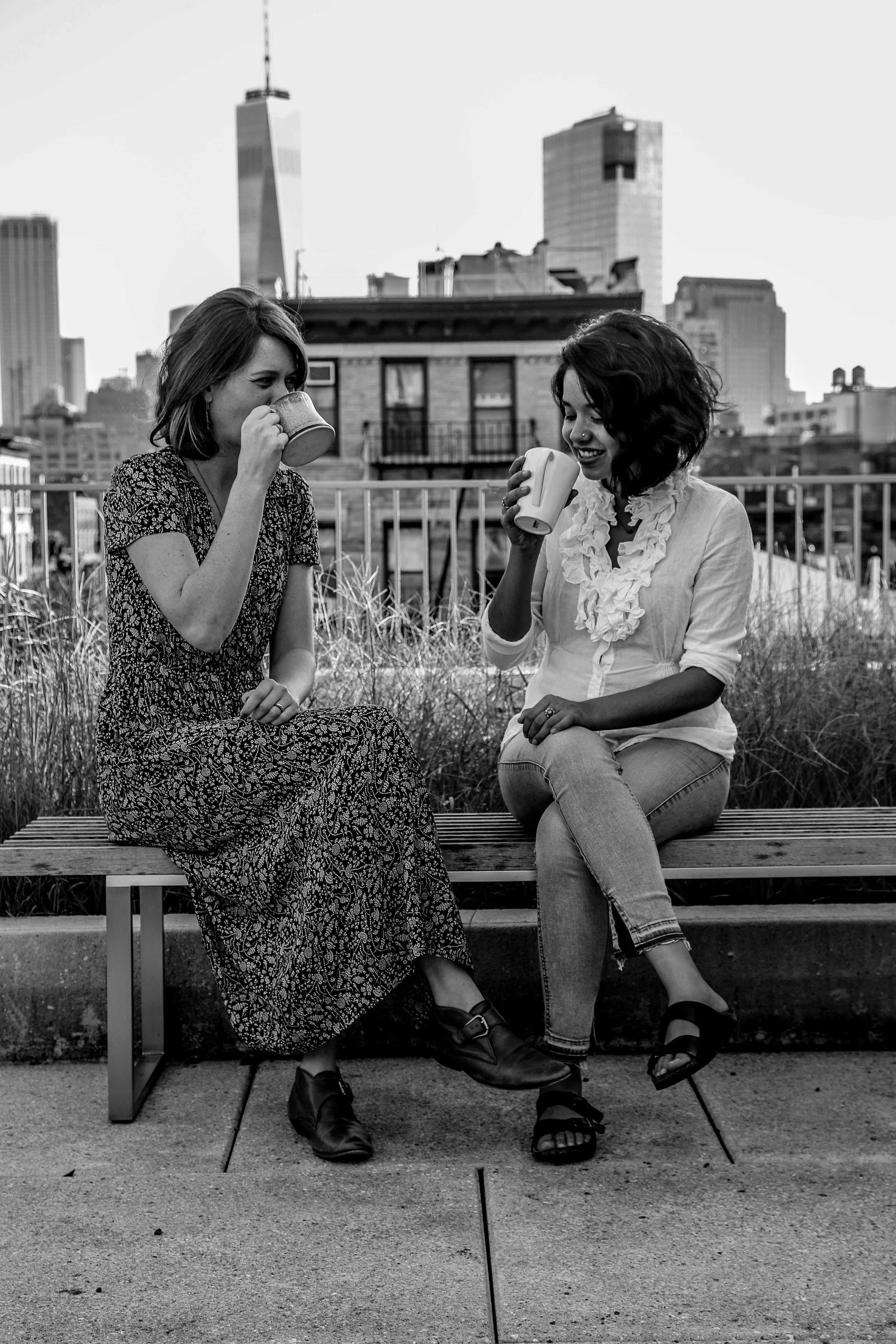 Anna Bulman and Ria Singh Sawhney sitting on a bench on a New York City rooftop drinking a cup of tea, engaged in a happy conversation