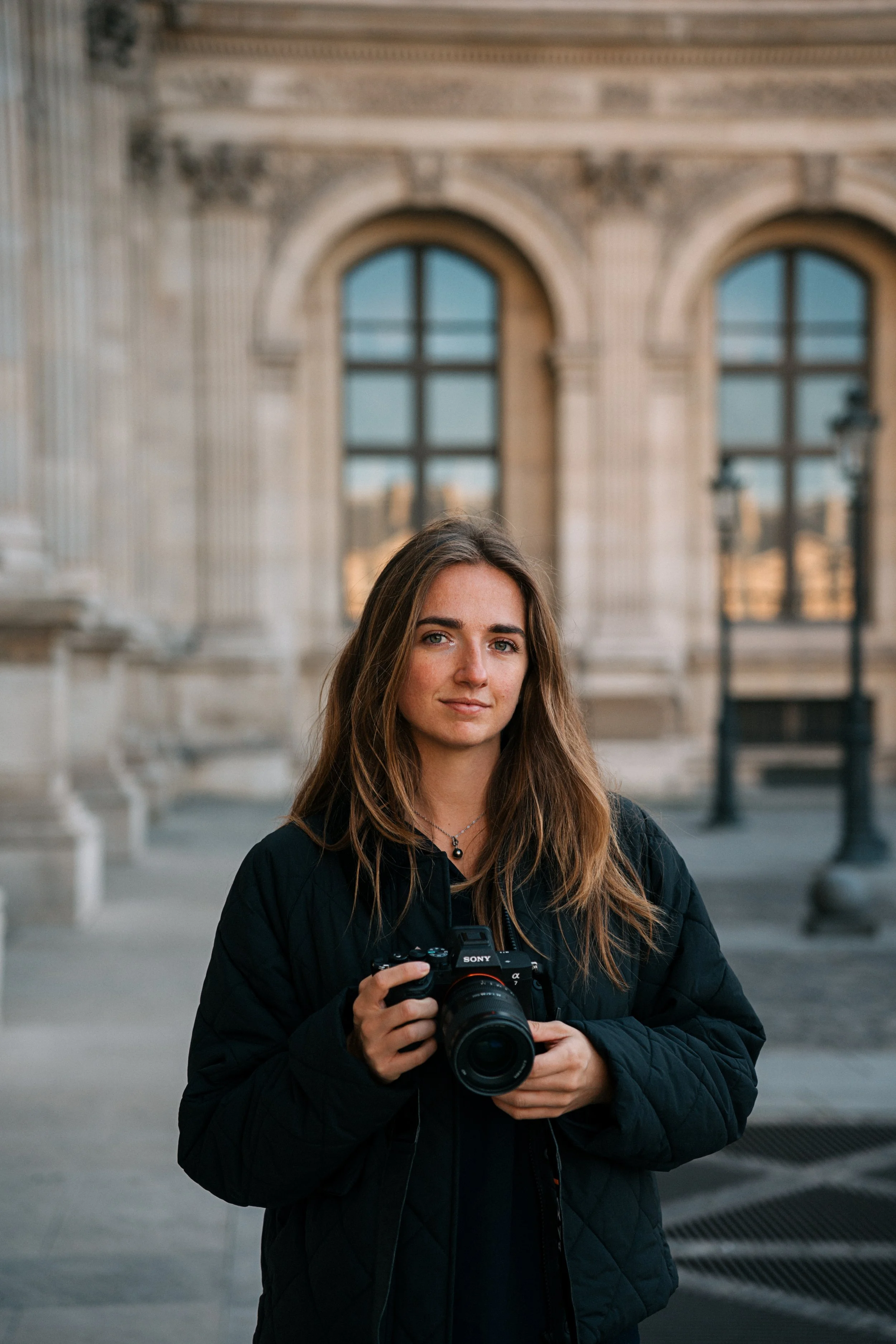 Une jeune femme avec des cheveux longs, portant un manteau noir et une chaîne, tenant un appareil photo Sony, se tient devant un bâtiment en pierre avec de grandes fenêtres et des lampadaires, en fin d'après-midi.