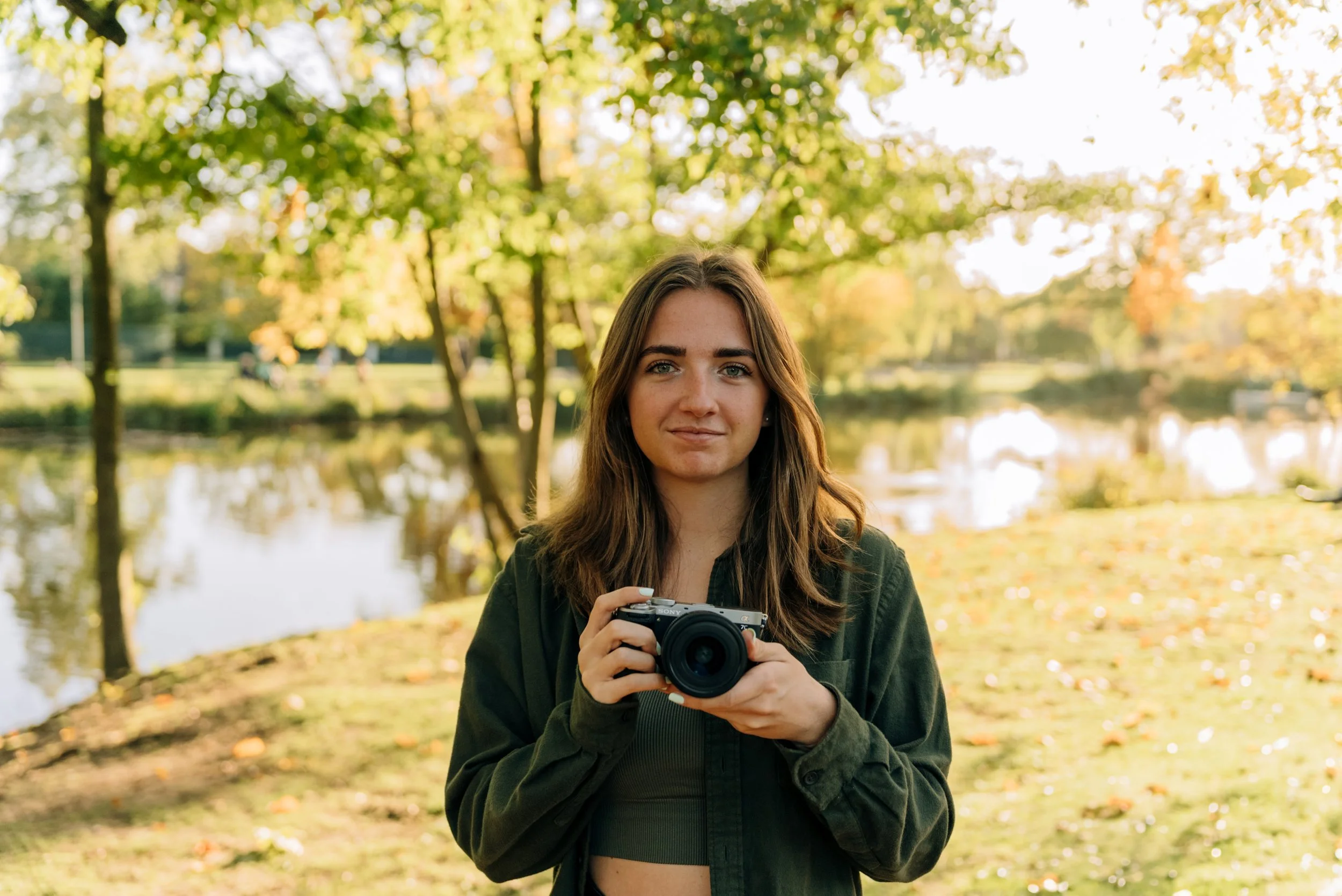 Une jeune femme souriante tenant un appareil photo dans un parc avec des arbres aux feuilles d'automne et un lac en arrière-plan.