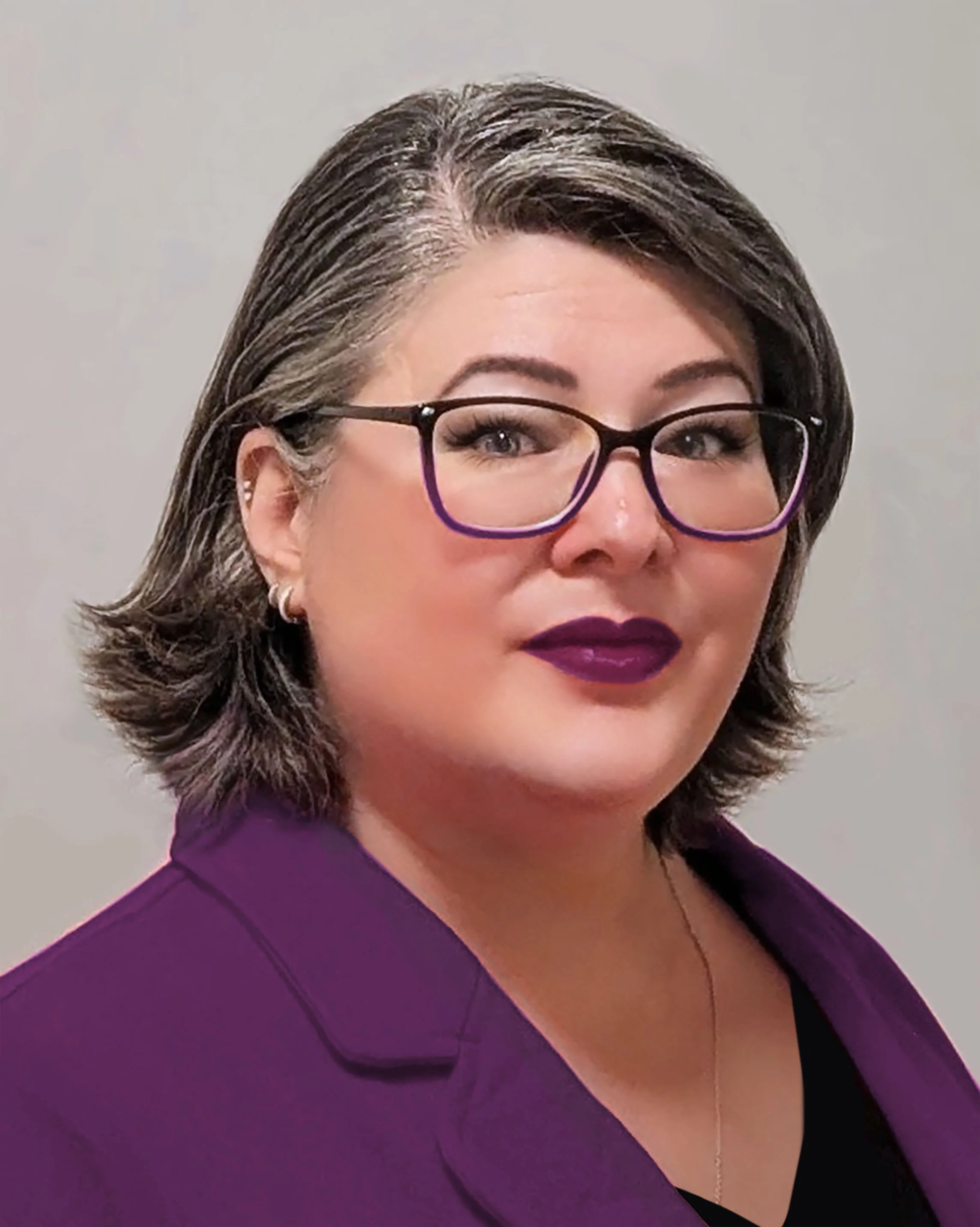 Image is a headshot of a woman with mid length salt and pepper hair  who wears glasses and smiles with a closed mouth. She is wearing a purple blazer.