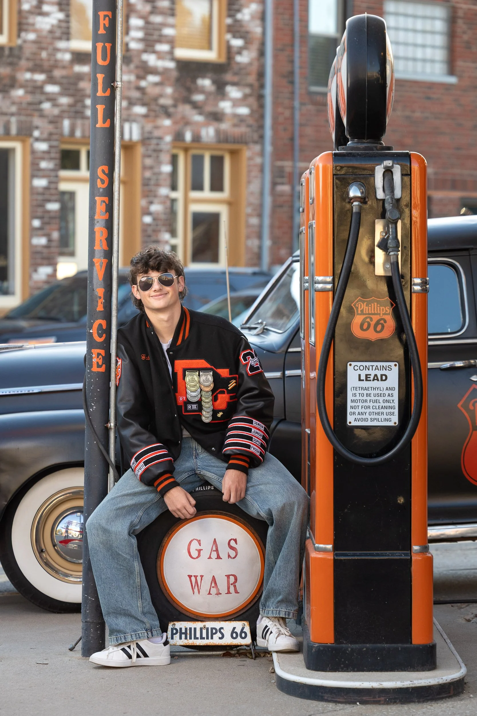 Senior portrait at a retro gas station featuring a teen in a patterned shirt leaning against a vintage orange Phillips 66 gas pump.