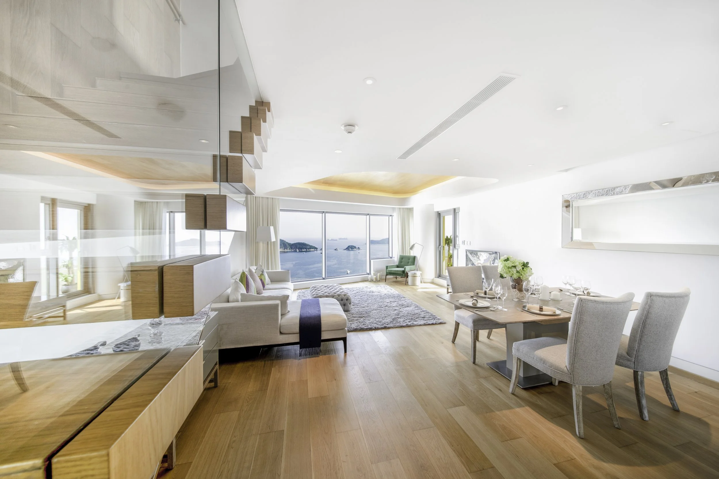 Bright, modern living room with large ocean view, white walls, wooden floors, white couch, green armchair, dining table with four chairs, decorative plants, and large windows.